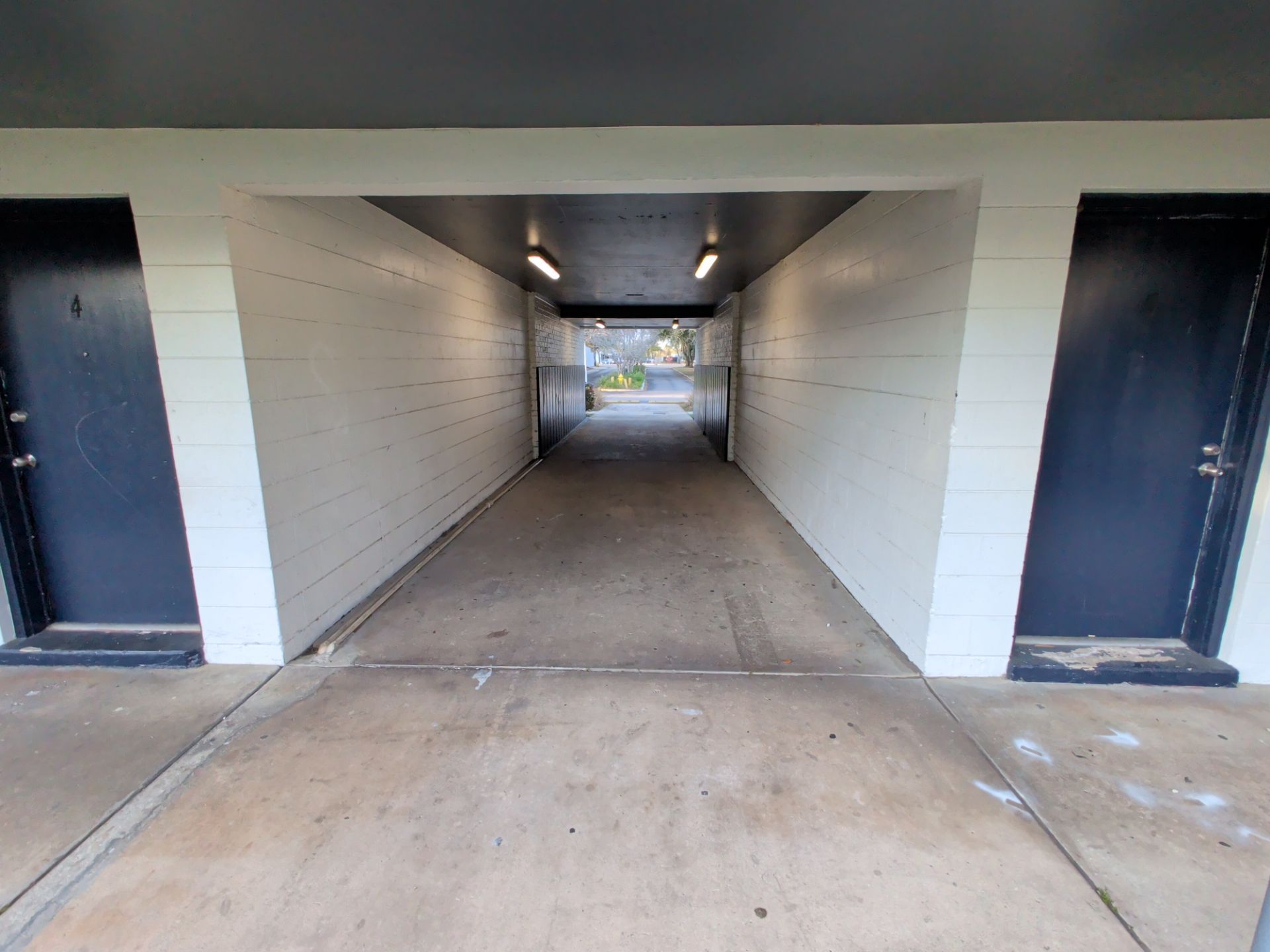 Narrow concrete passageway with white walls and dark doors leading to a lit outdoor area