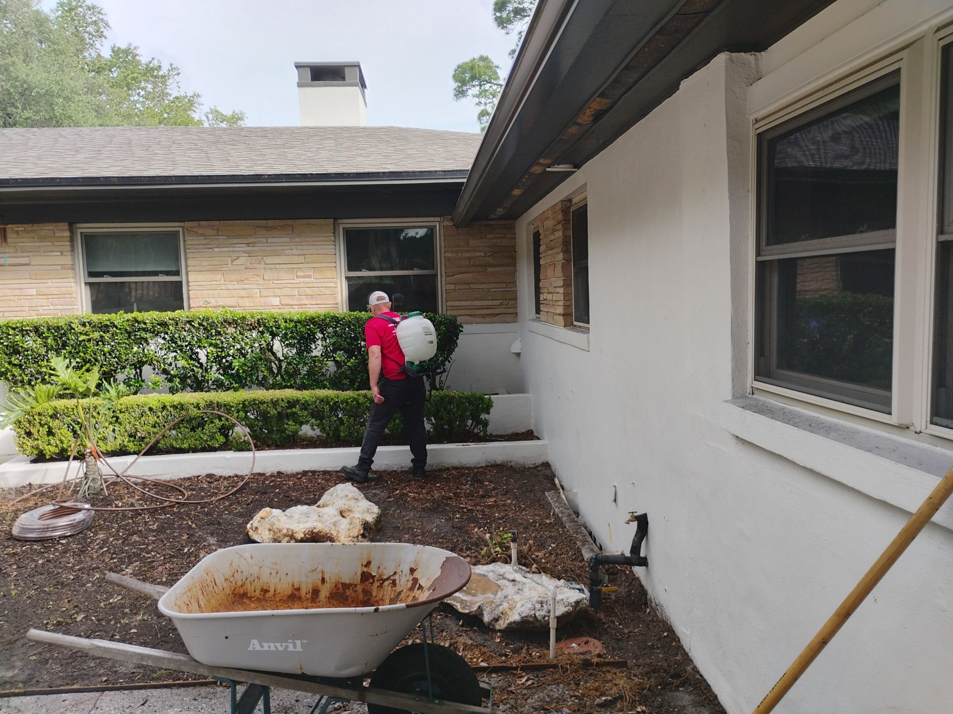 Person in red backpack near a house exterior, with a wheelbarrow, mulch, and landscaping supplies.