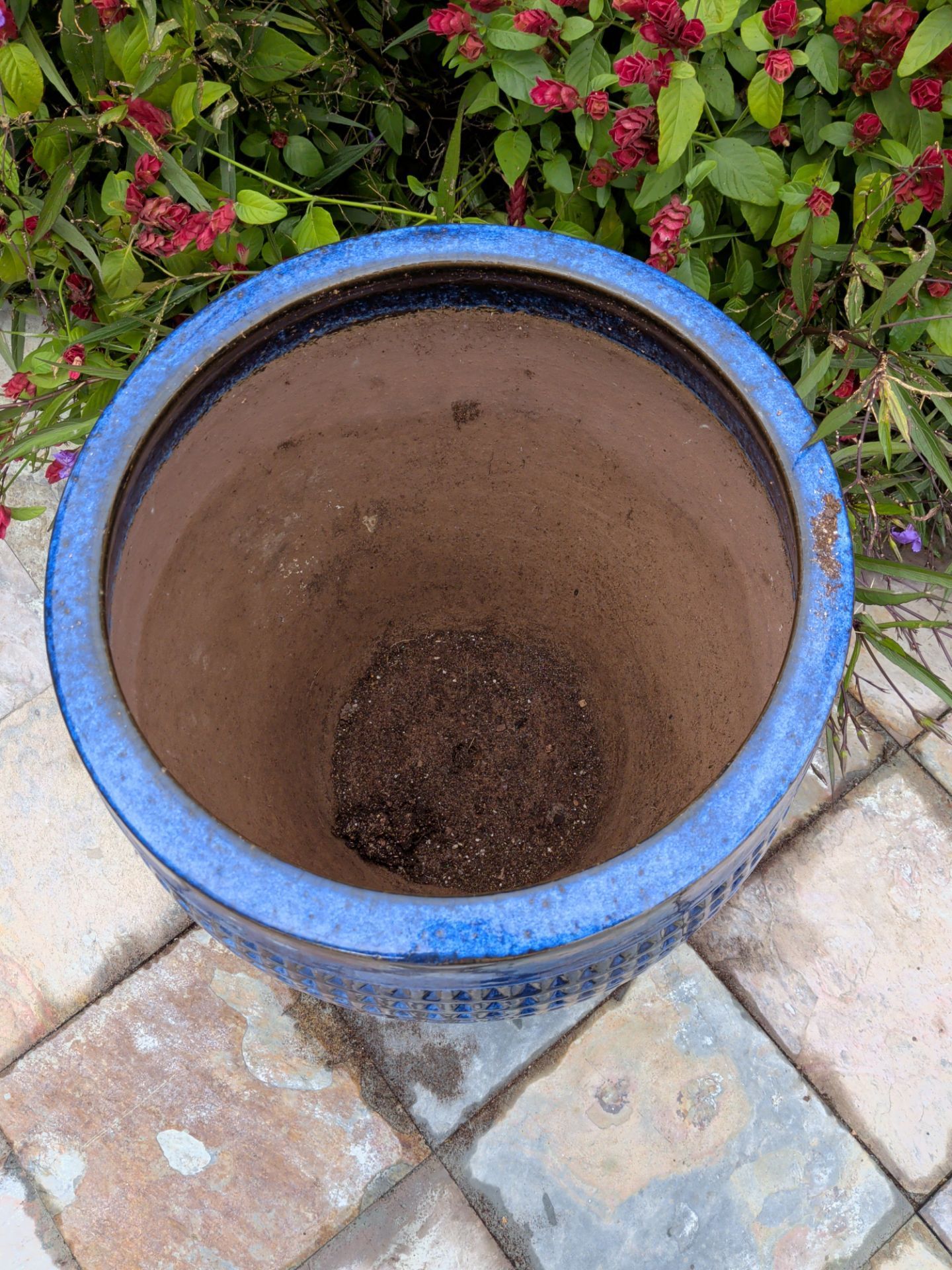 Blue ceramic planter pot with soil, sitting on a tiled patio beside pink flowers.