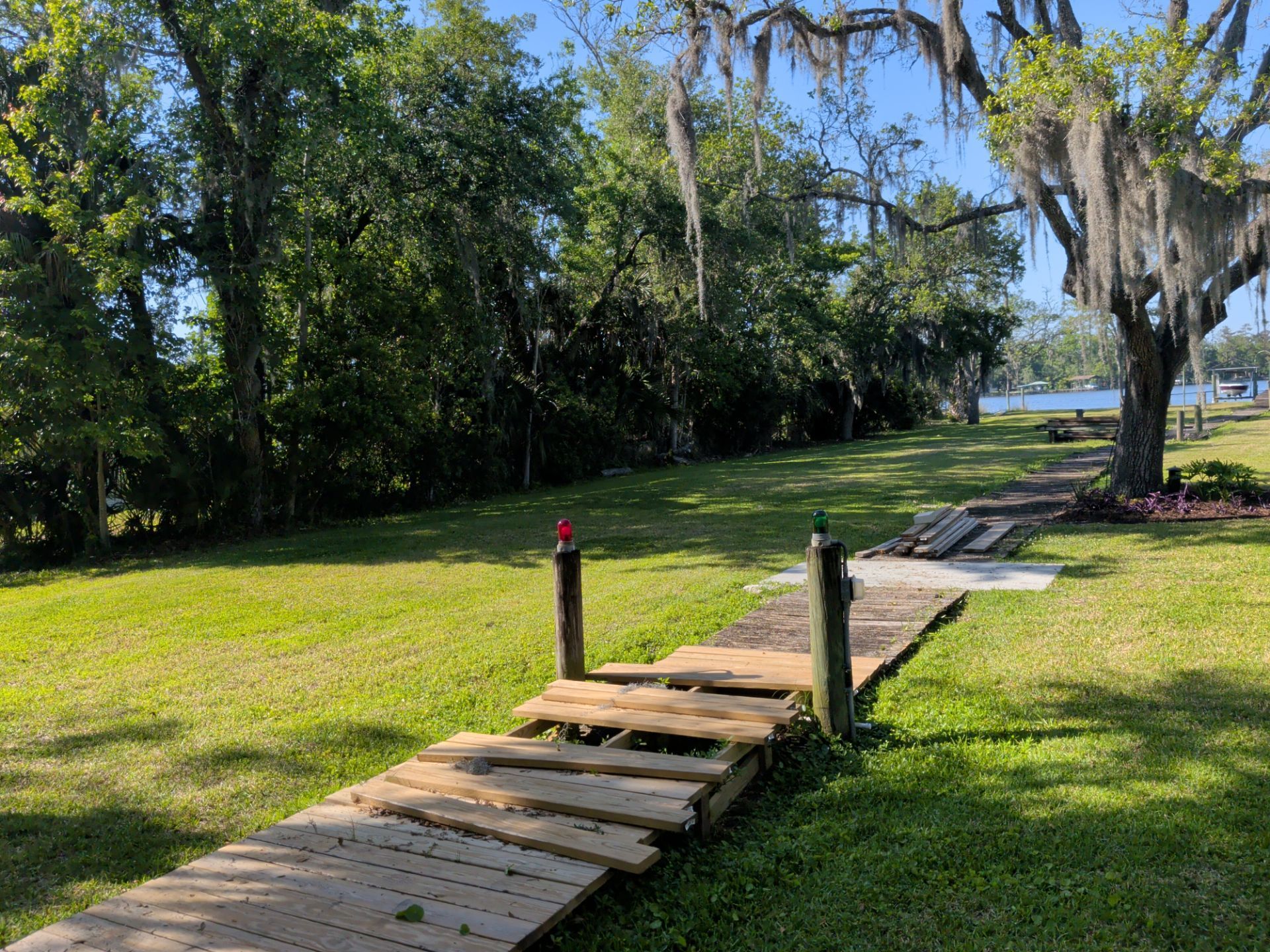 Wooden boardwalk through grassy park leading to a waterfront under trees and blue sky