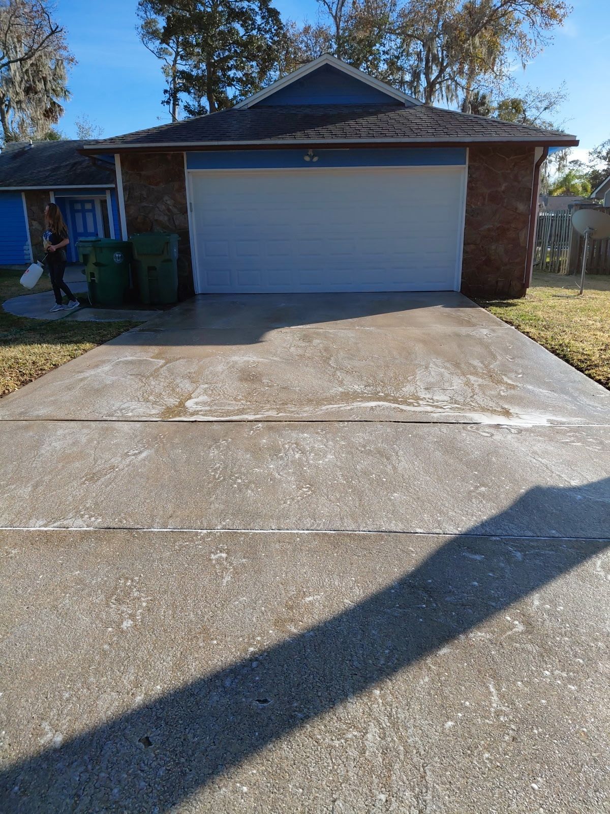 Driveway of a house with a closed garage door. Person stands near the trash cans holding a bucket.