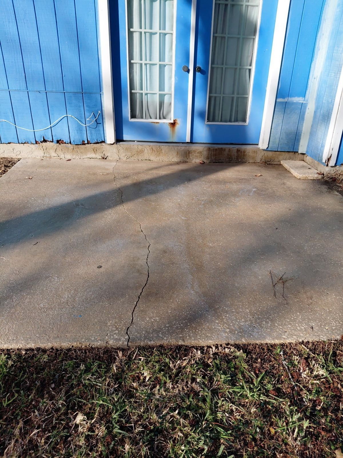 Concrete patio with a crack, in front of a blue doorway with white curtains.