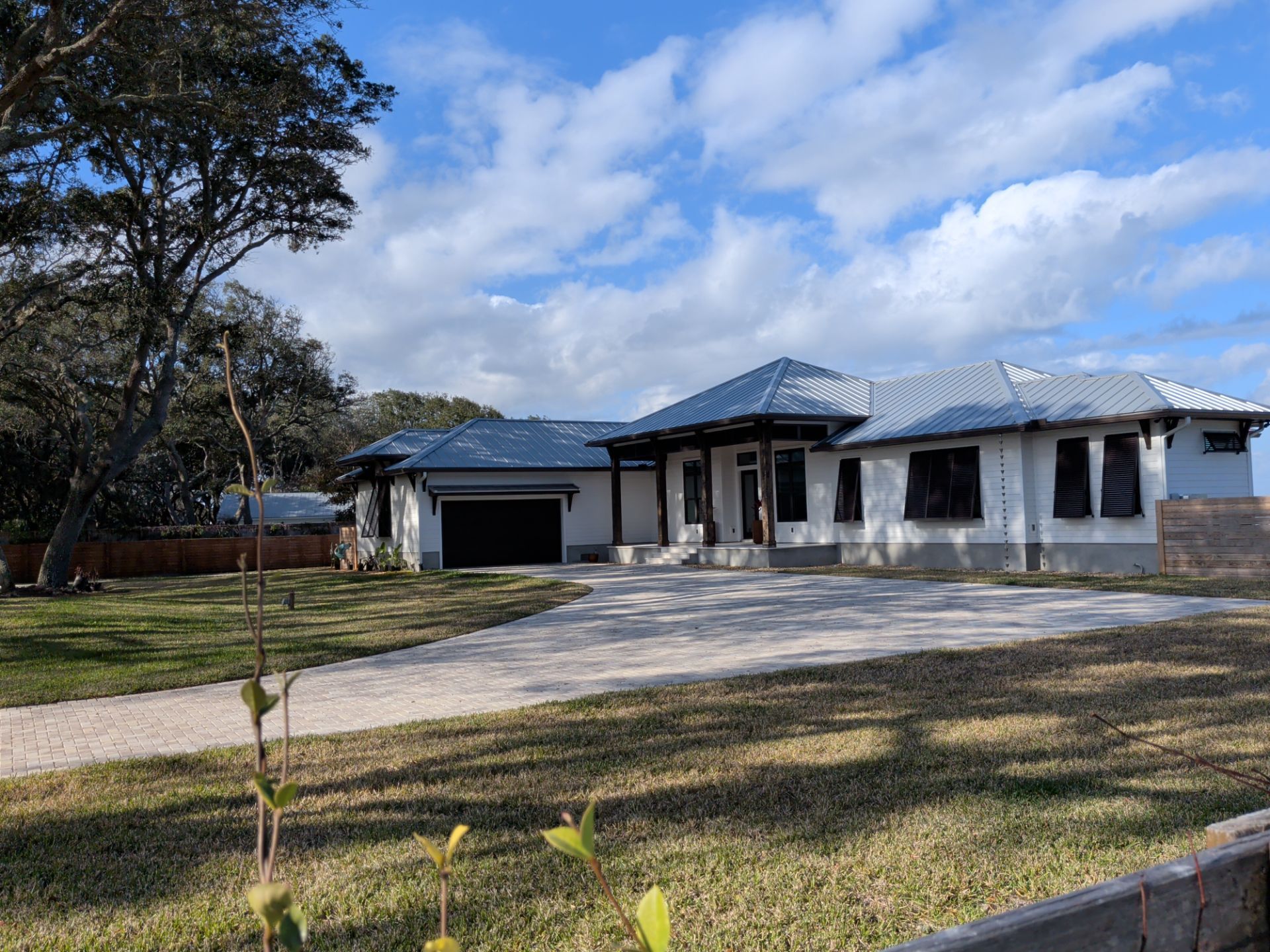 Single-story white house with a metal roof, driveway, and large tree under a blue sky