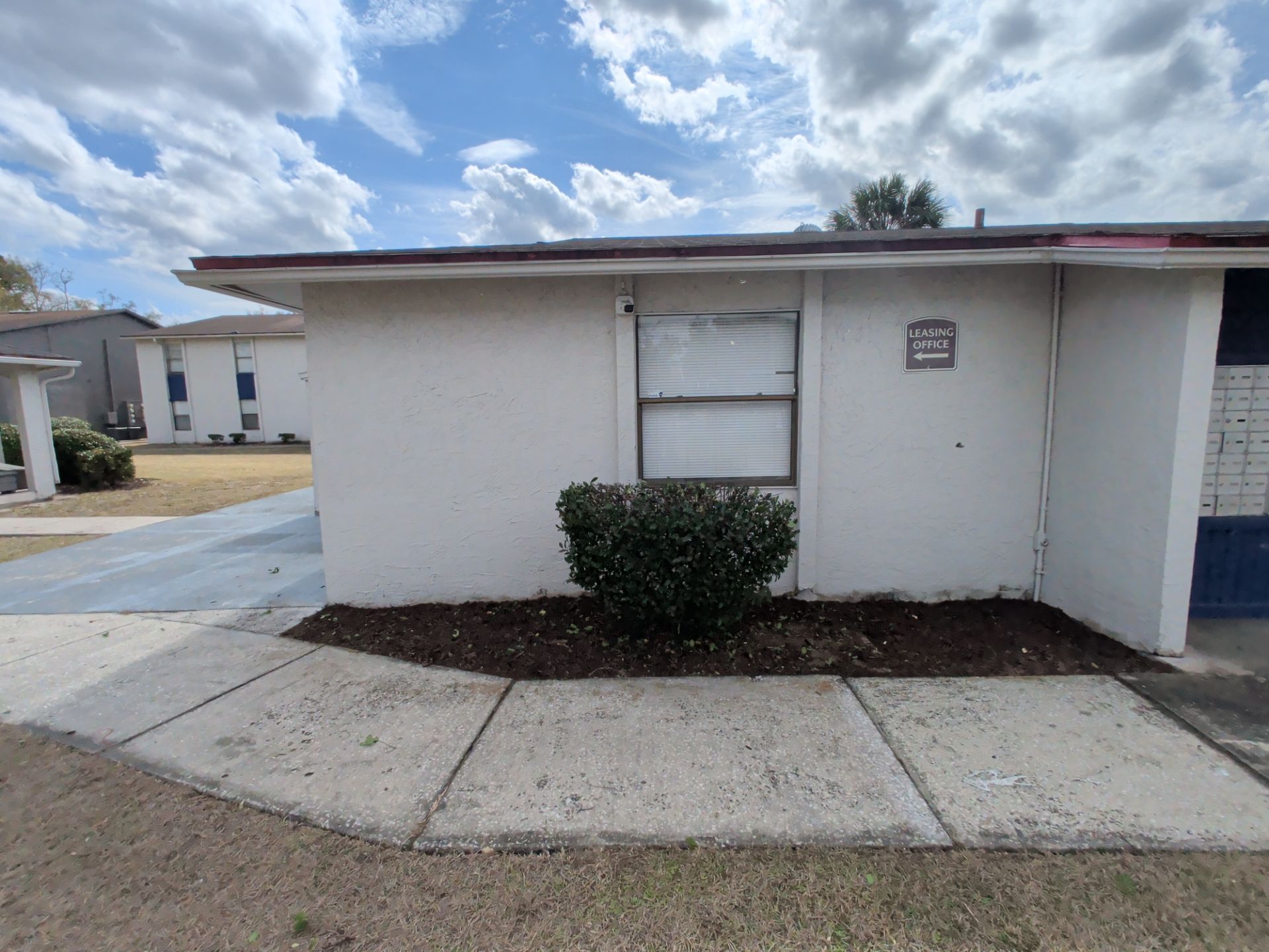 Small white utility building with a door, bush, and concrete walkway under a cloudy sky