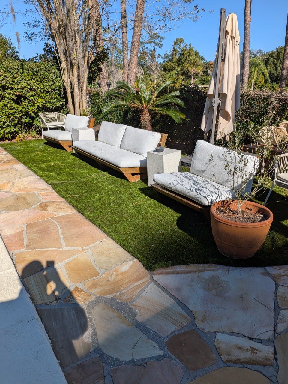 Outdoor patio with wicker sofa, patterned bench, umbrella, potted plant, and stone walkway beside green lawn