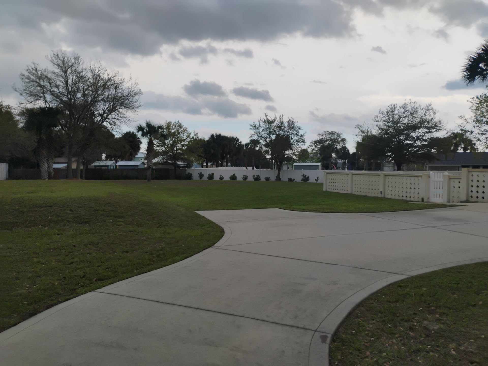 Curving concrete path through a grassy park under a cloudy sky, with trees and white fences in the background