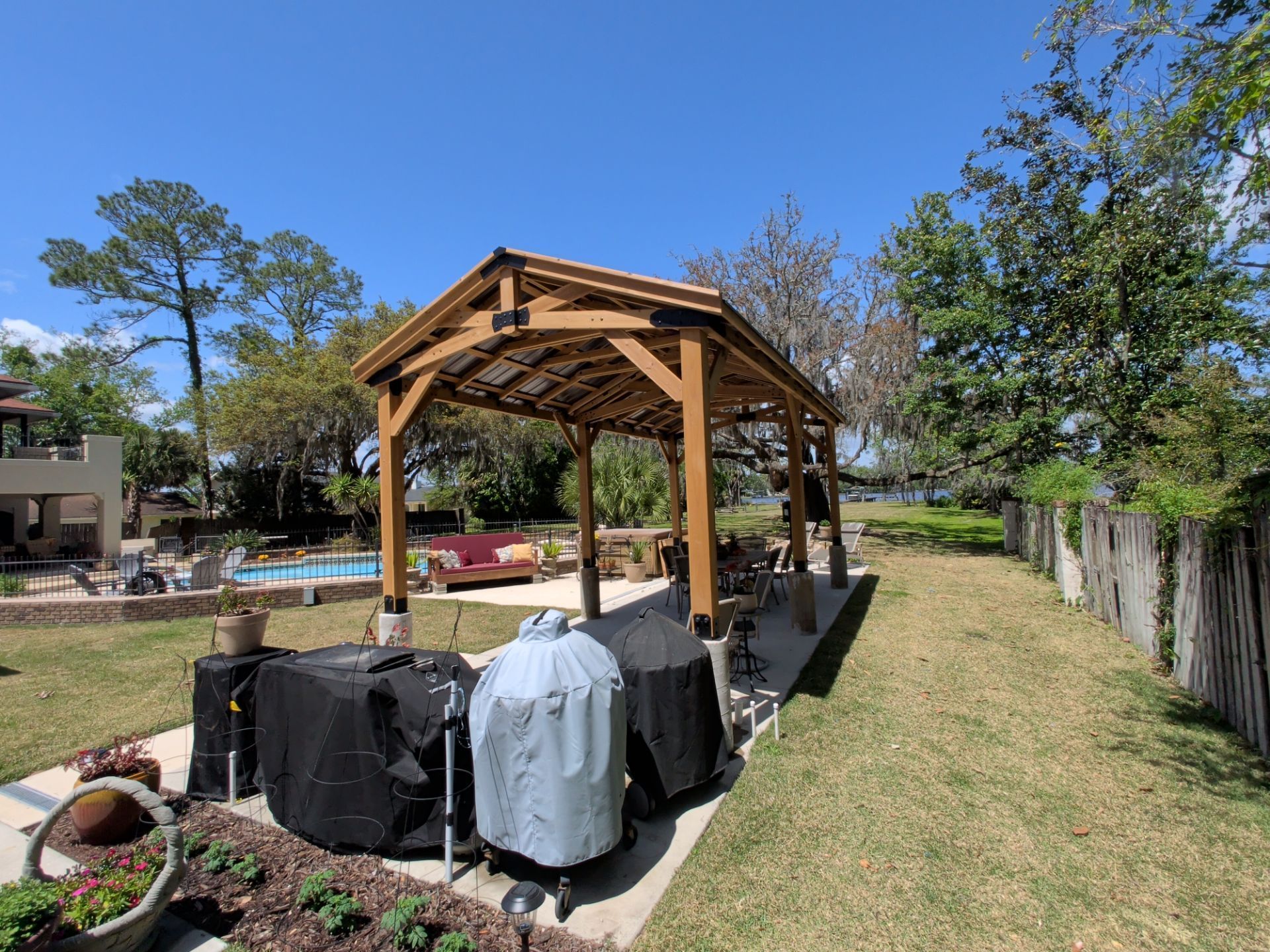 Wooden gazebo on a grassy backyard patio with covered grills and a clear blue sky