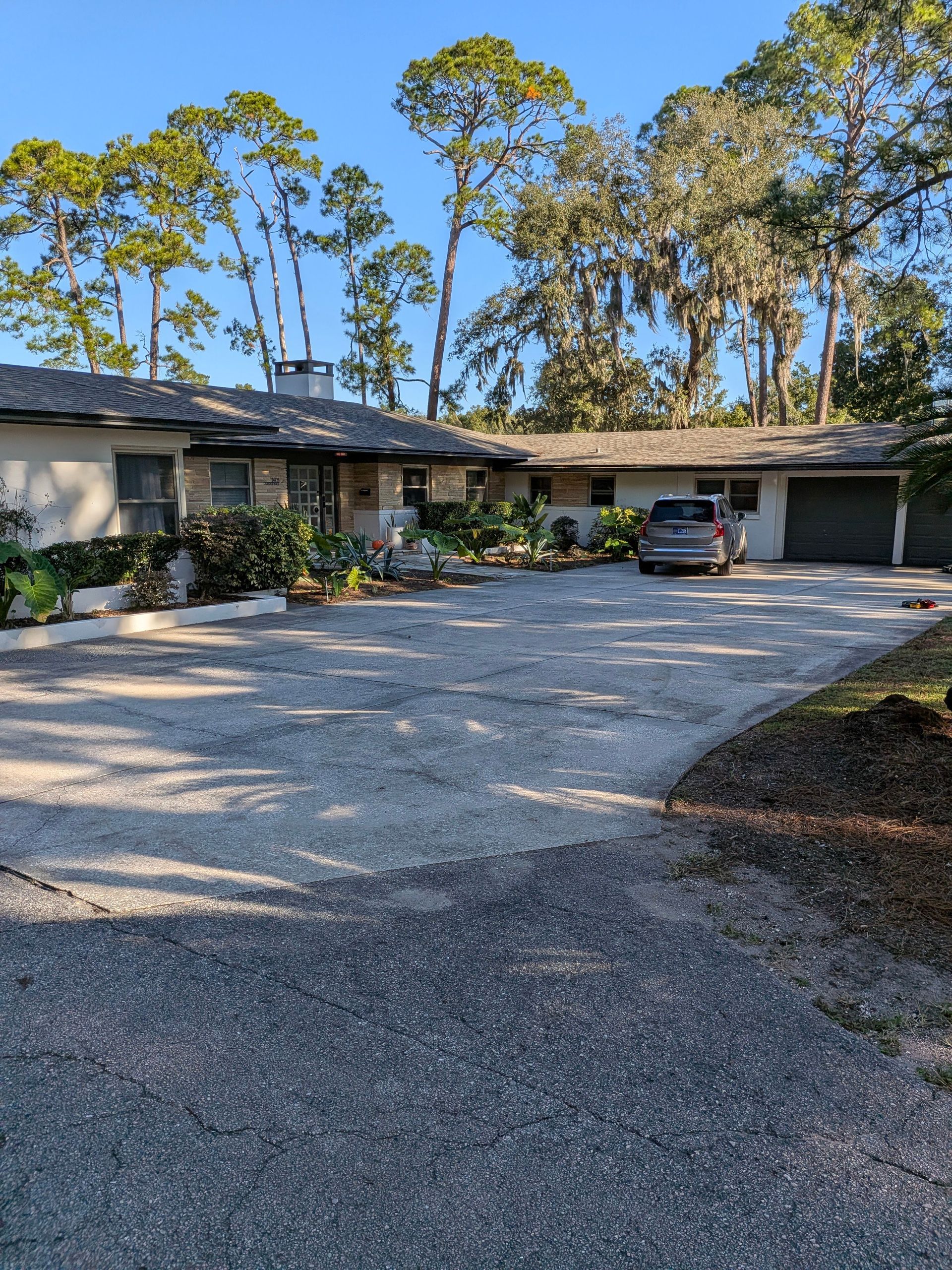 Single-story house with a gravel driveway, parked SUV, and tall trees in the background.