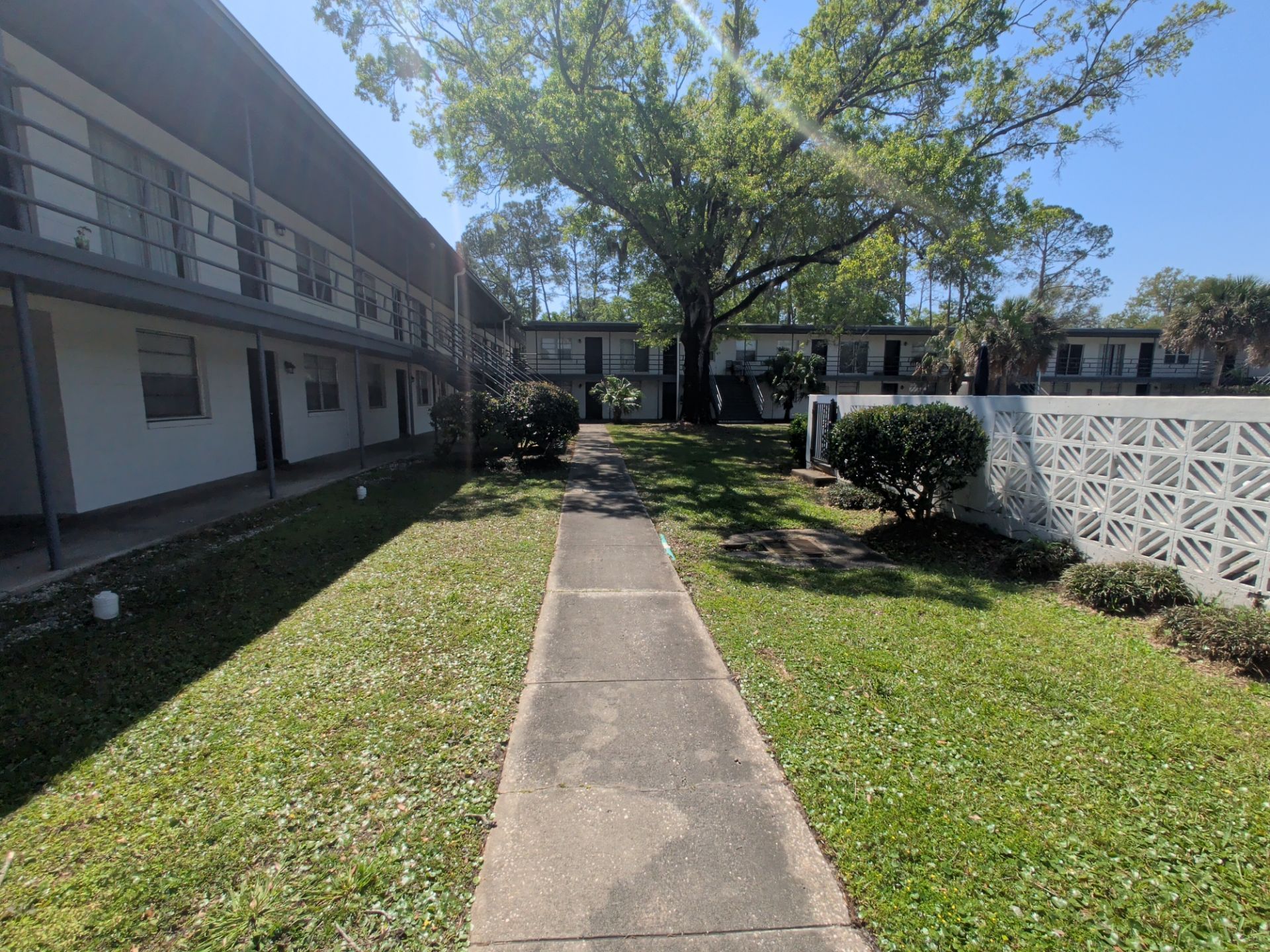 Walkway through grassy courtyard beside a white apartment building and fence, with trees and shrubs.