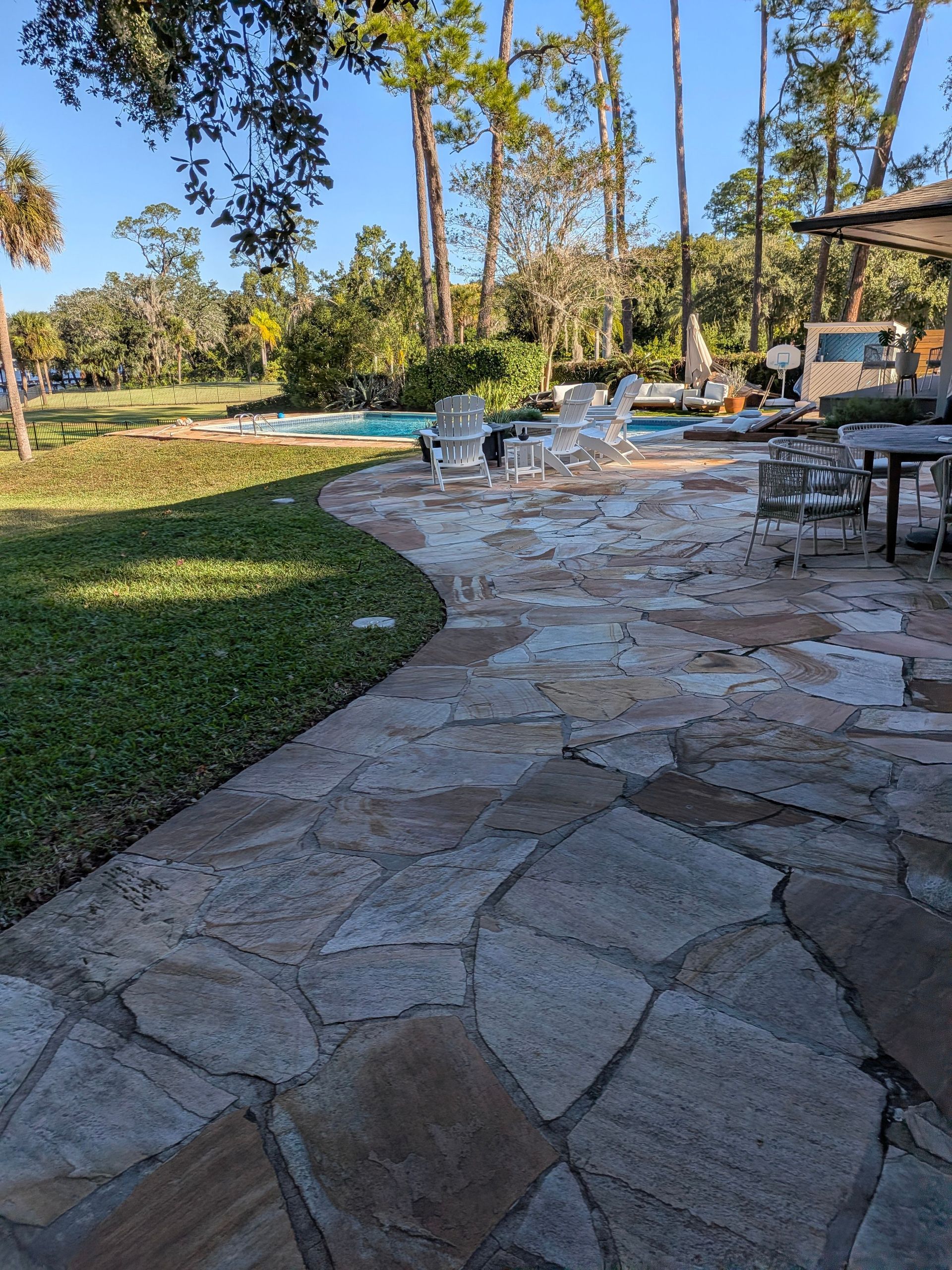 Stone patio with outdoor chairs beside a pool, trees, and a grassy yard in sunlight