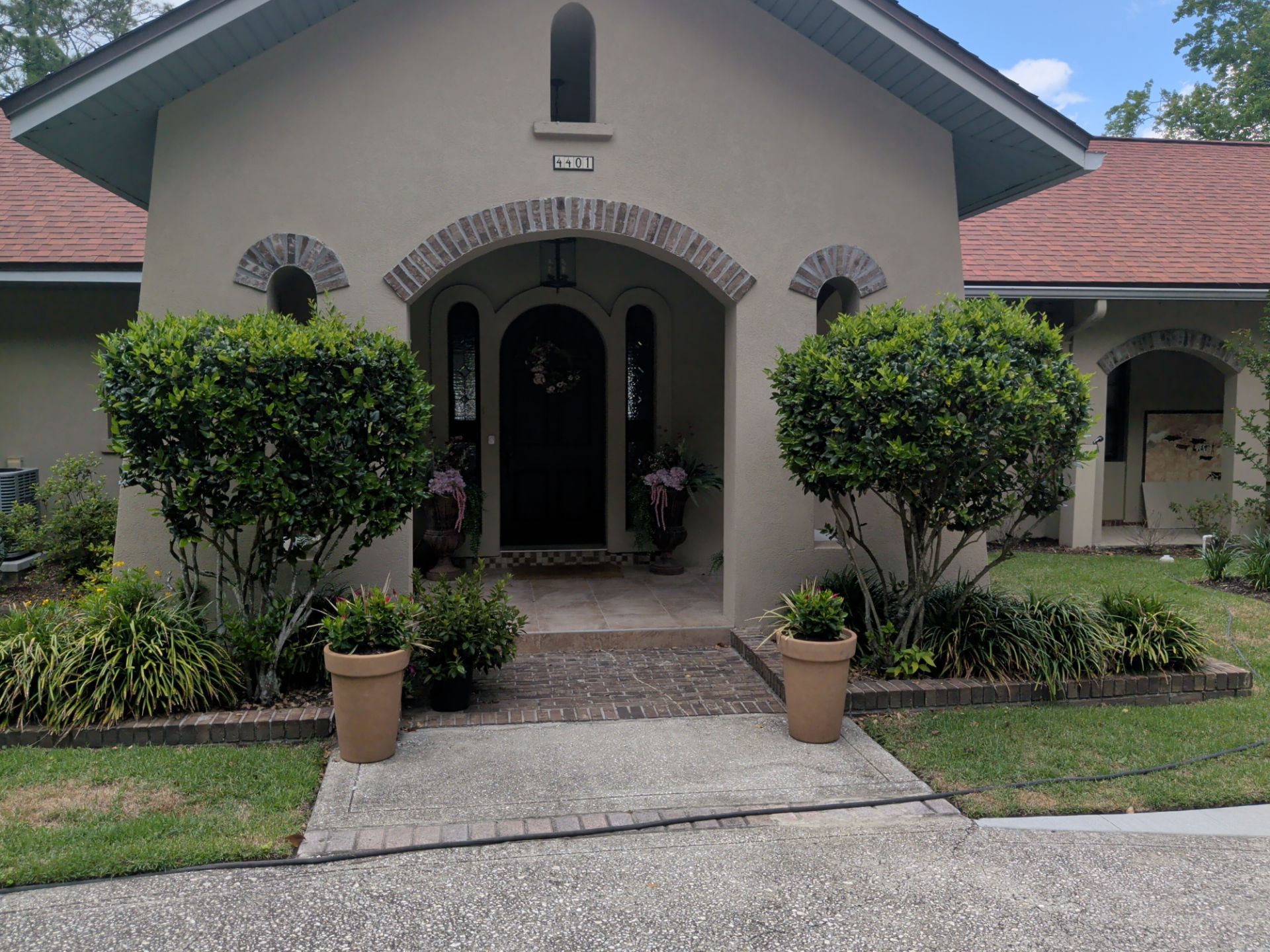 Front view of a house with arched entry, trimmed shrubs, and potted plants along a walkway