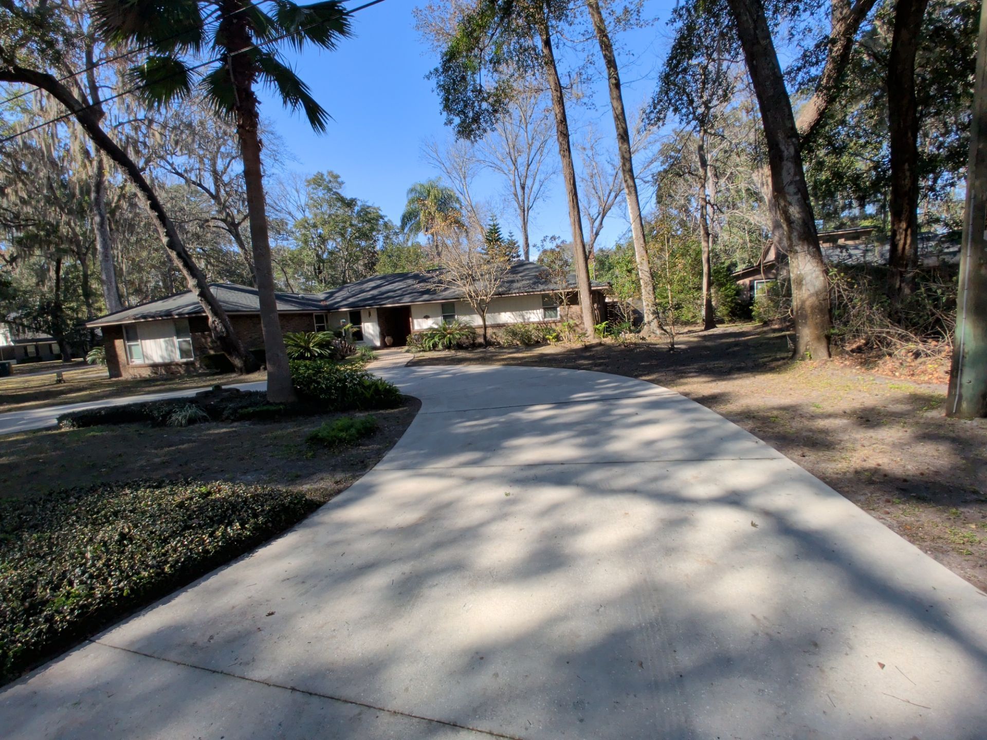 A long concrete driveway leading to a single-story home surrounded by trees and greenery under a clear, blue sky.