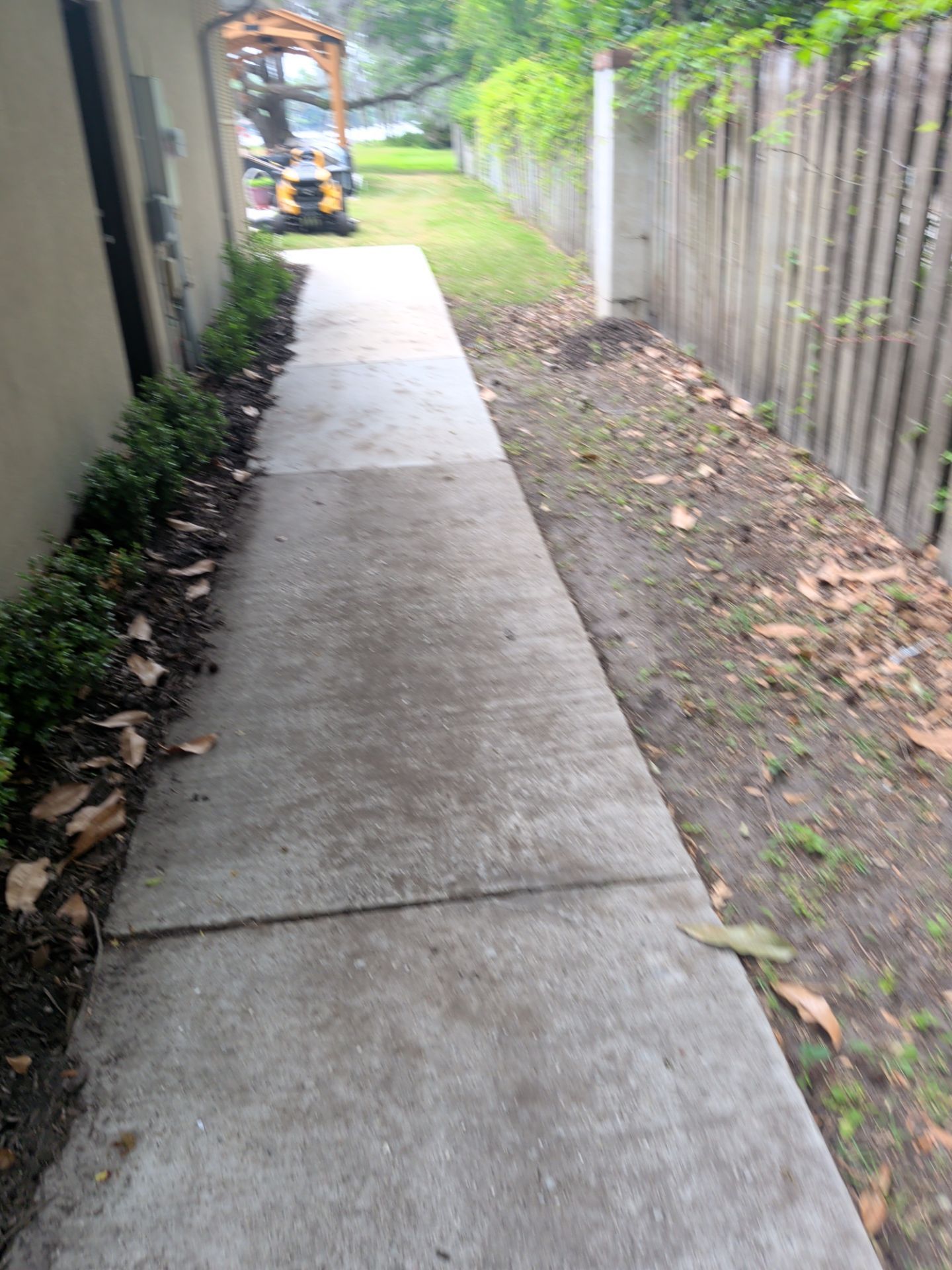 Narrow concrete walkway between a house and wooden fence, leading to a backyard patio area.