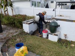 Person spraying around a grill and outdoor kitchen.  White fence and shrubbery in the background.