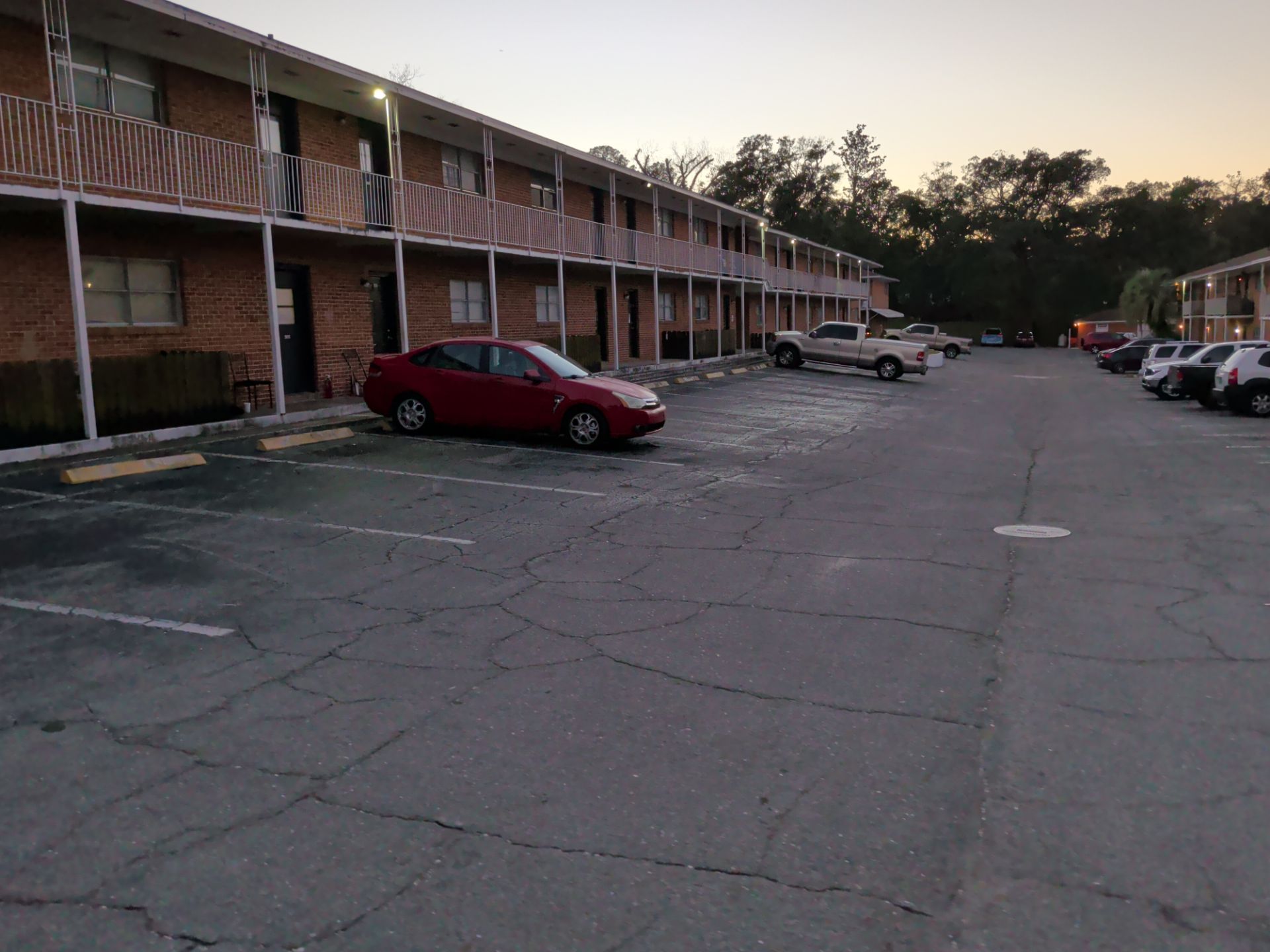 Apartment parking lot with a red car and several parked vehicles at dusk.