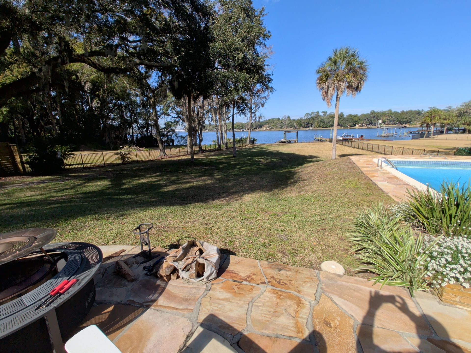 Lawn and pool area overlooking a body of water with trees, blue sky, and a palm tree.