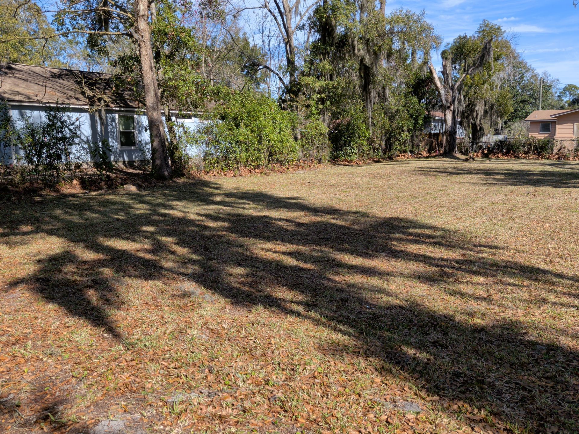 A cleared grassy lot with long shadows cast by trees, bordered by a fence and some buildings.