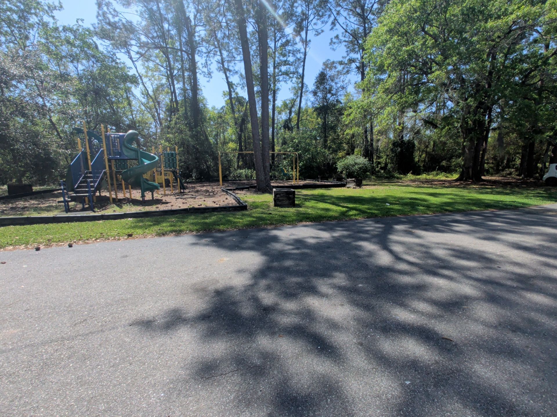 Sunny park with playground equipment, trees, and a paved path beside a grassy area