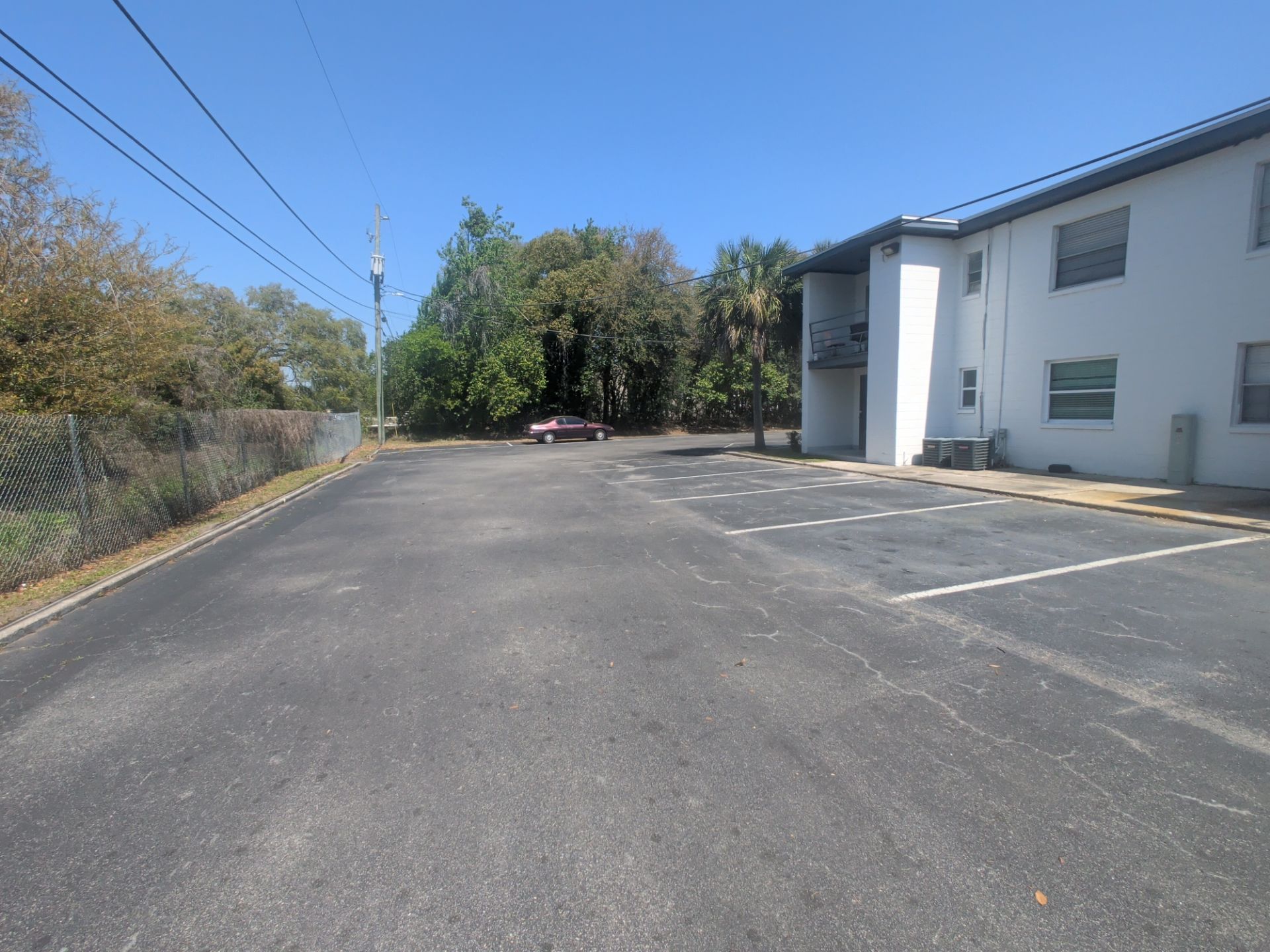 Empty asphalt parking lot beside a white building and chain-link fence under a clear blue sky