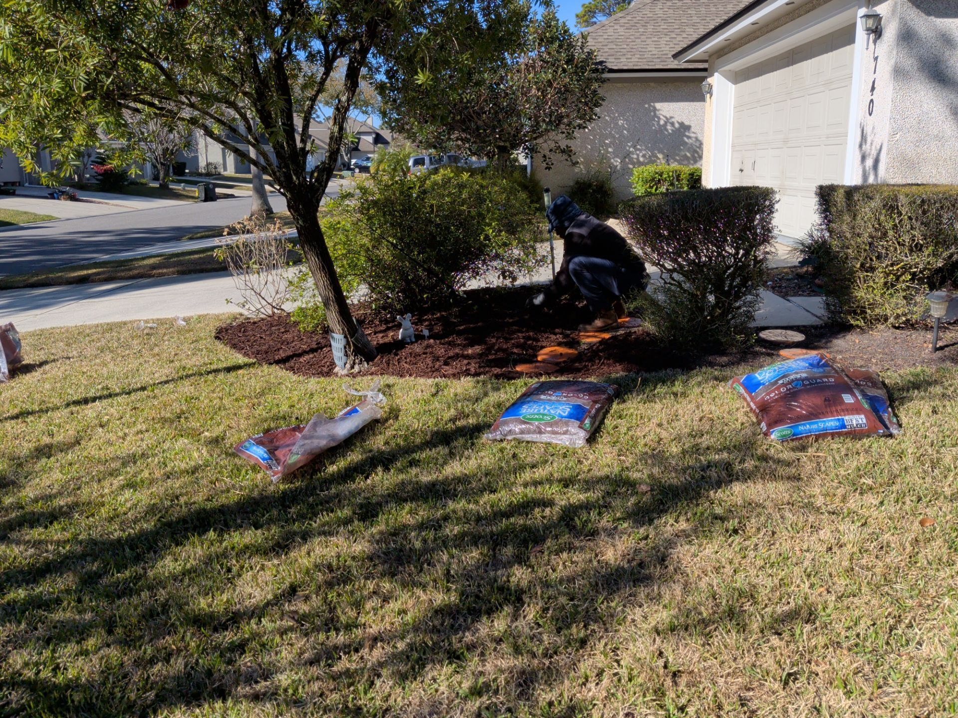 Person mulching a flower bed in front of a house, surrounded by bags of mulch on the grass.