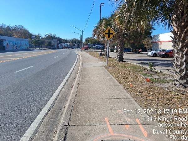 Wide suburban road with sidewalk, palm trees, and a yellow curve warning sign under clear blue sky