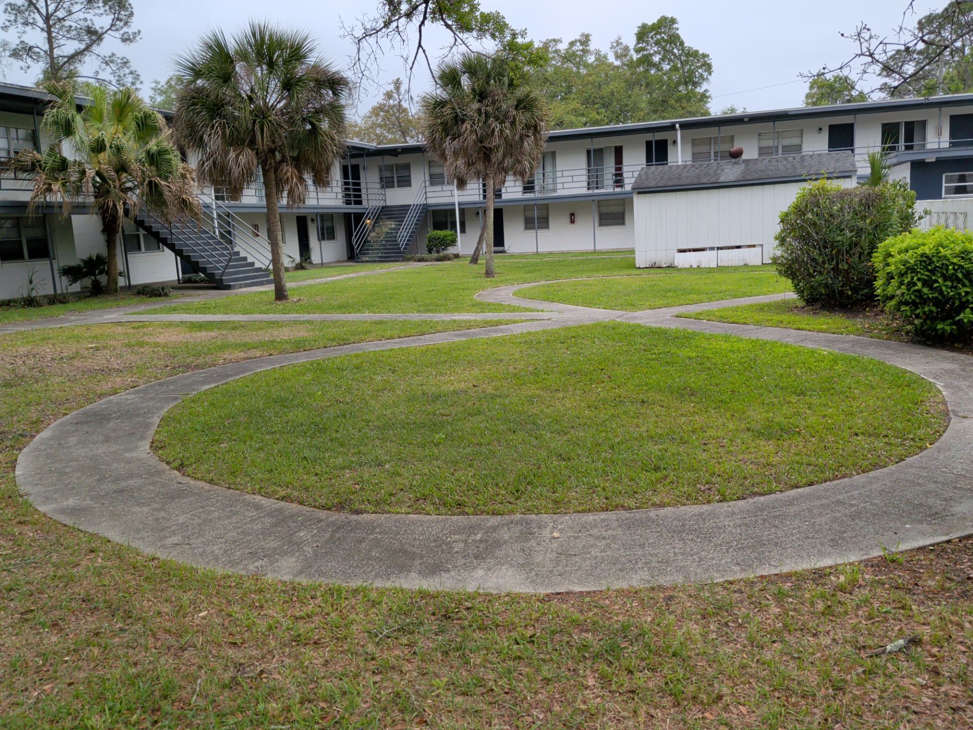 Courtyard with a circular concrete walkway around a grassy center, bordered by apartment buildings and palm trees.