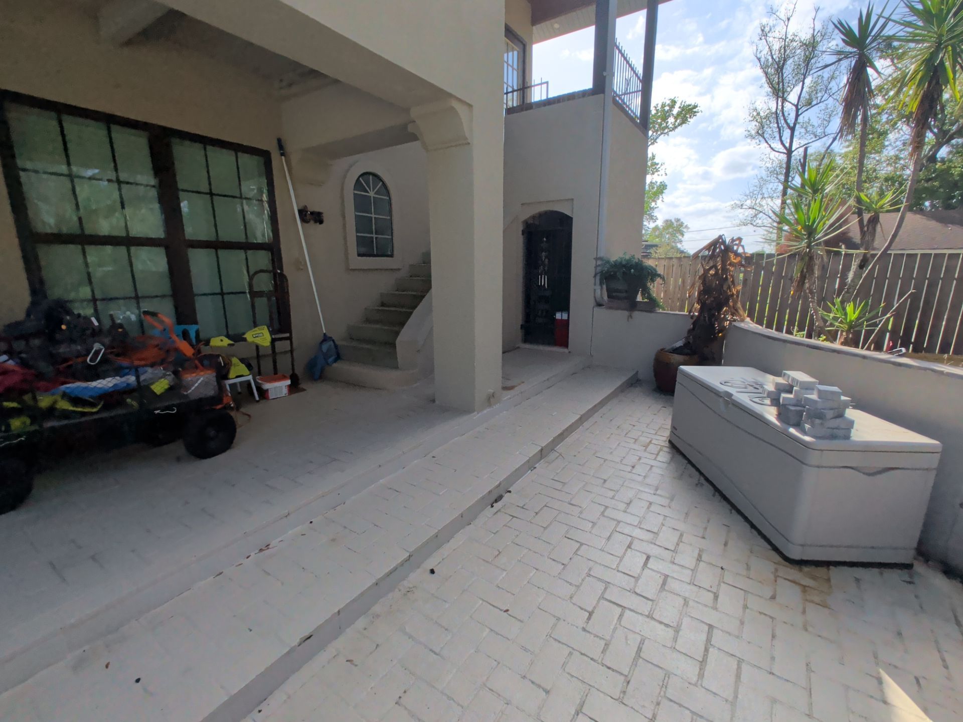 Covered patio with tiled floor, black-framed windows, stairs, and a gray outdoor sofa by a wooden fence.
