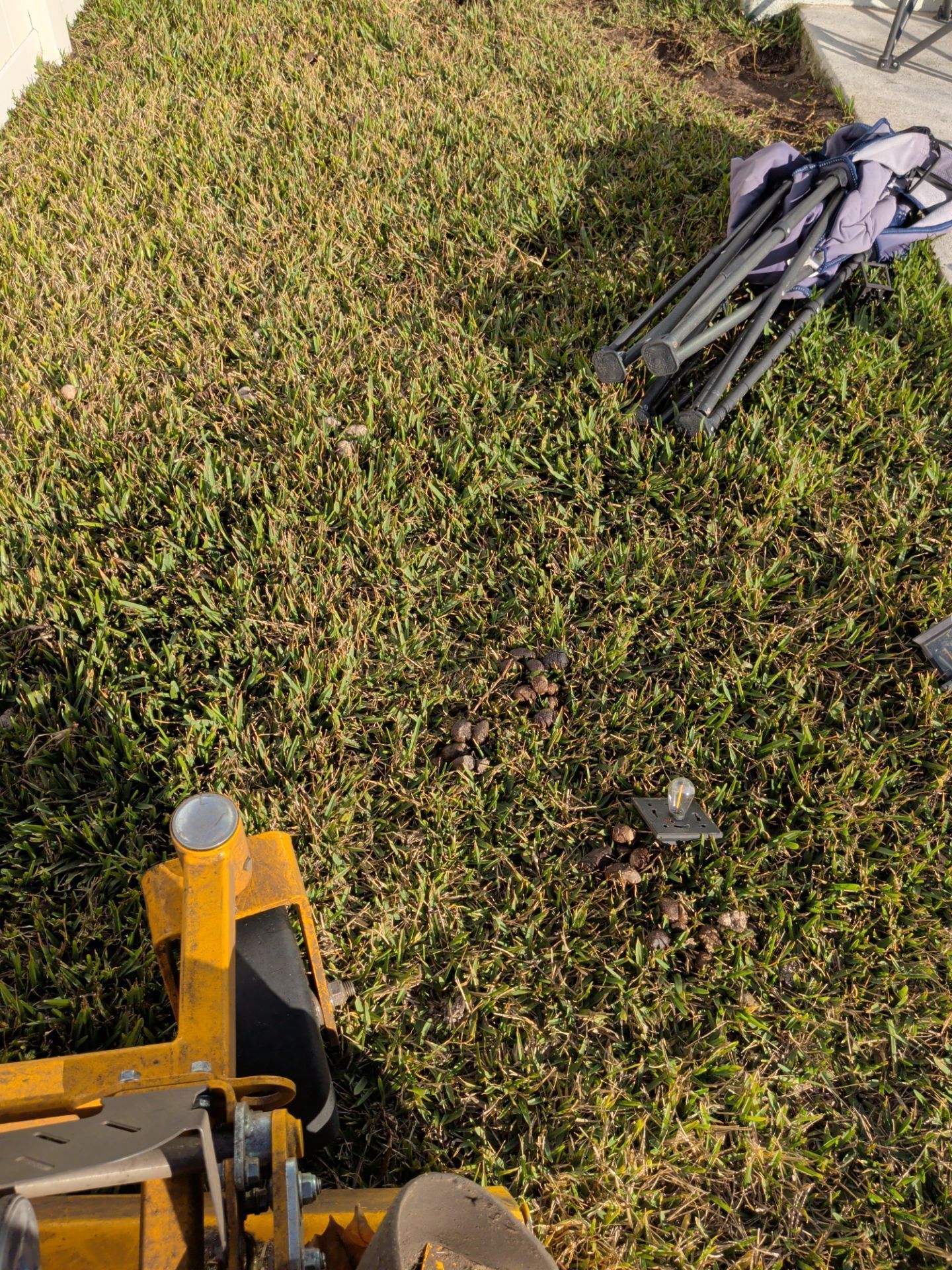 Yellow lawn mower cutting green grass with a folded gray tripod on the right side