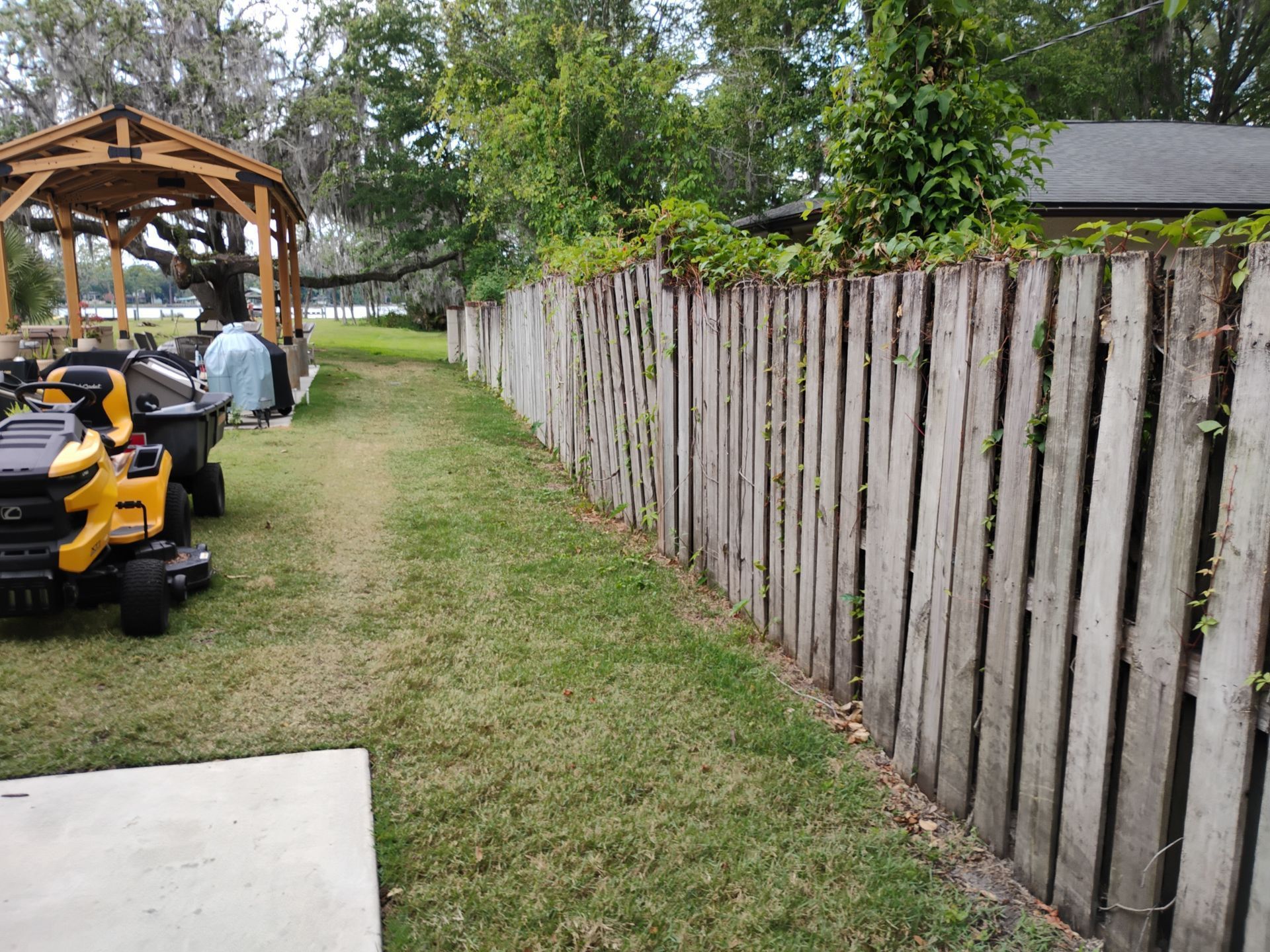 Backyard with wooden fence, grass path, gazebo, and a yellow lawn mower near a person.