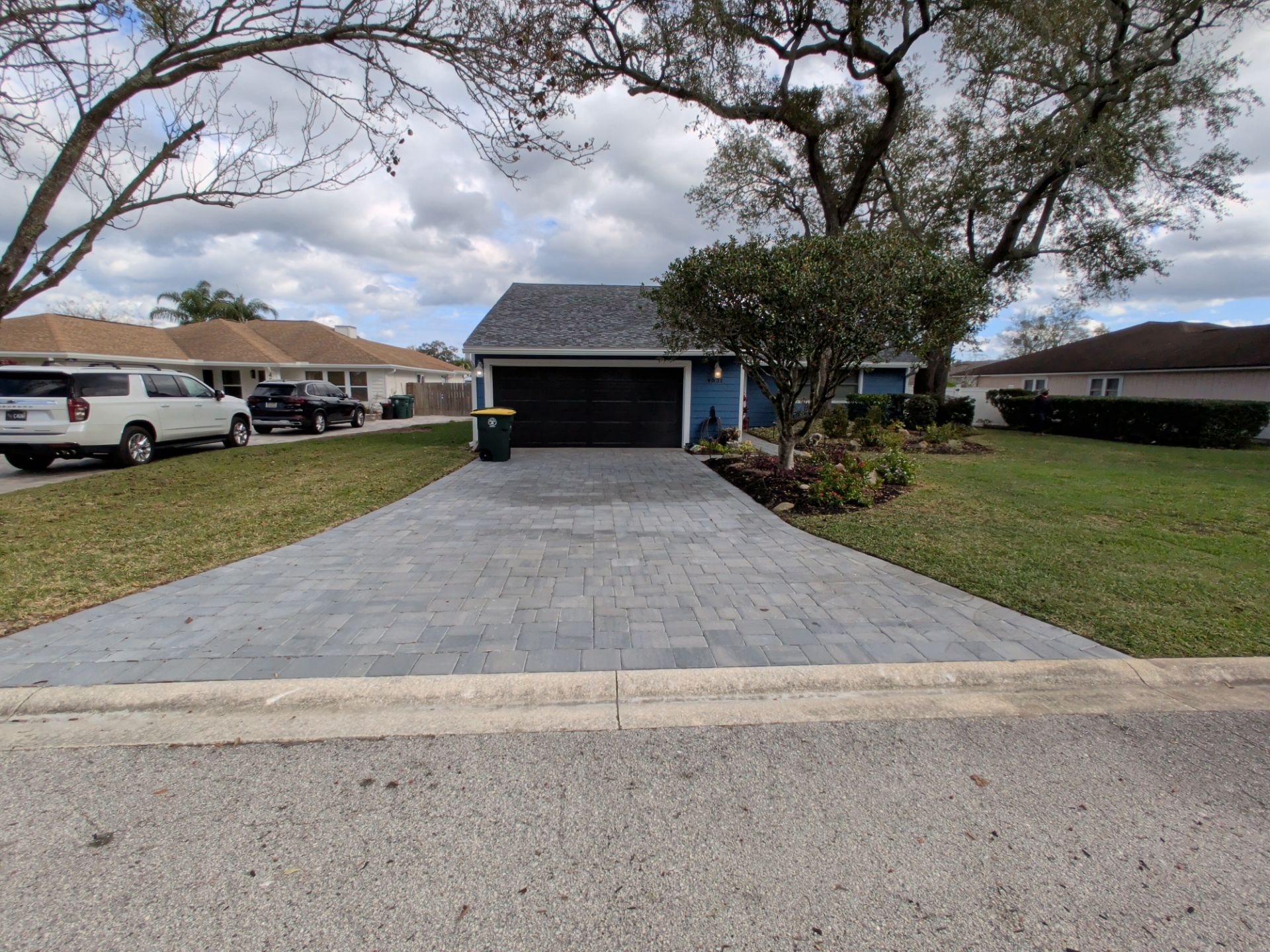 Blue house with gray paver driveway. Green lawn, trees, and parked cars on a cloudy day.