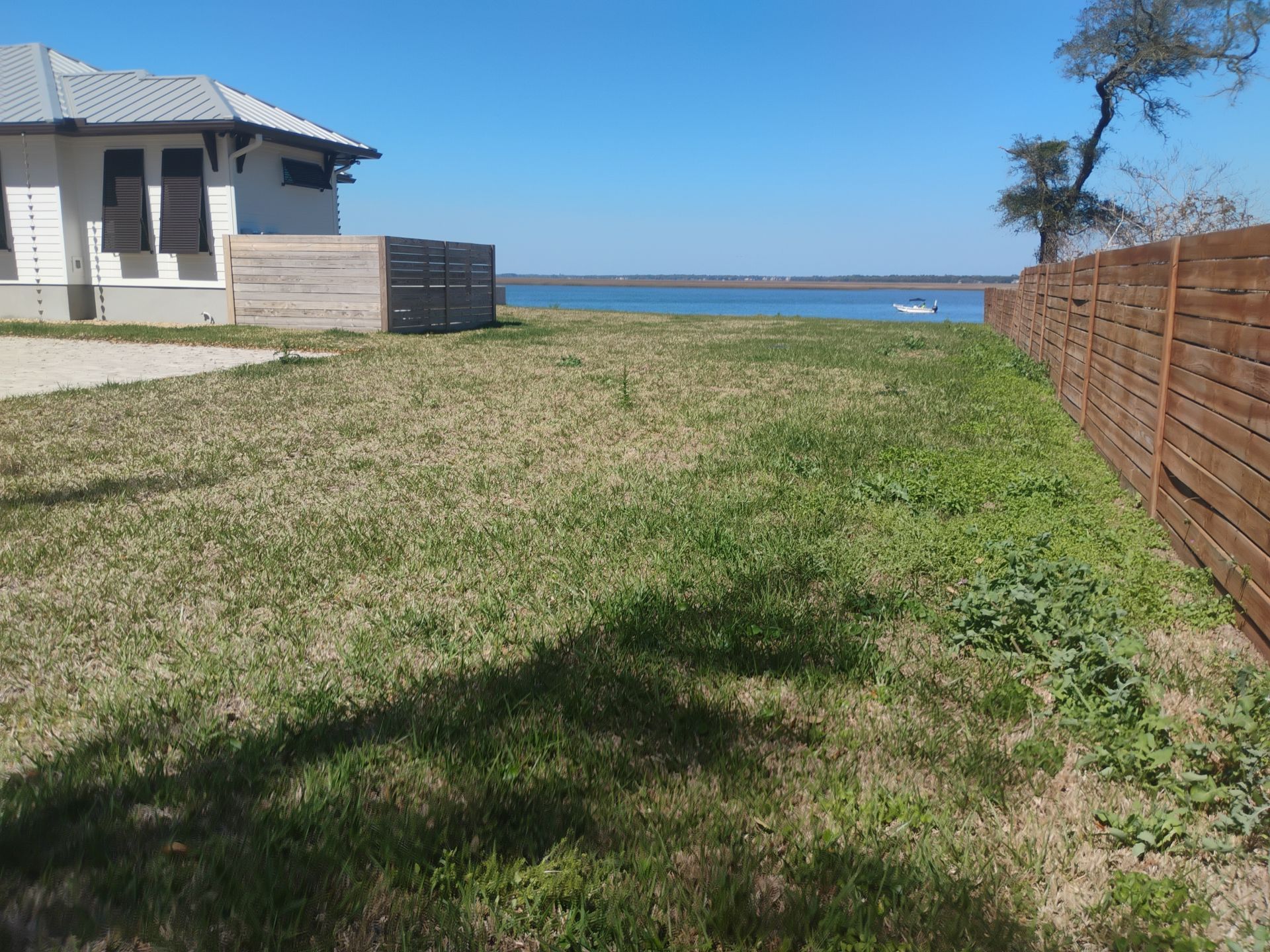 Grassy backyard leading to a blue lake, beside a house and wooden fence under a clear sky