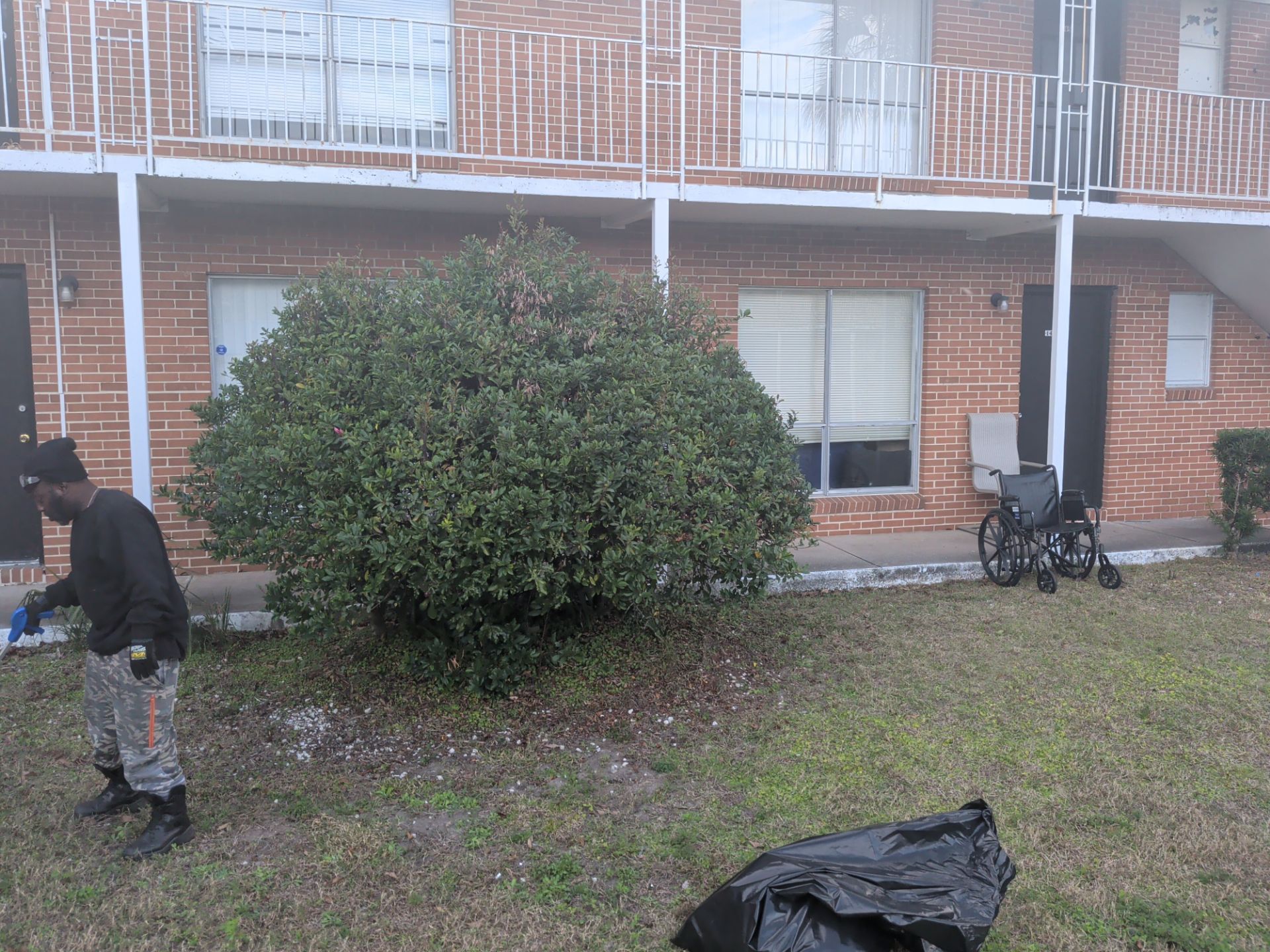 Person cleaning up yard by brick building, wheelchair nearby, trash bag on ground.
