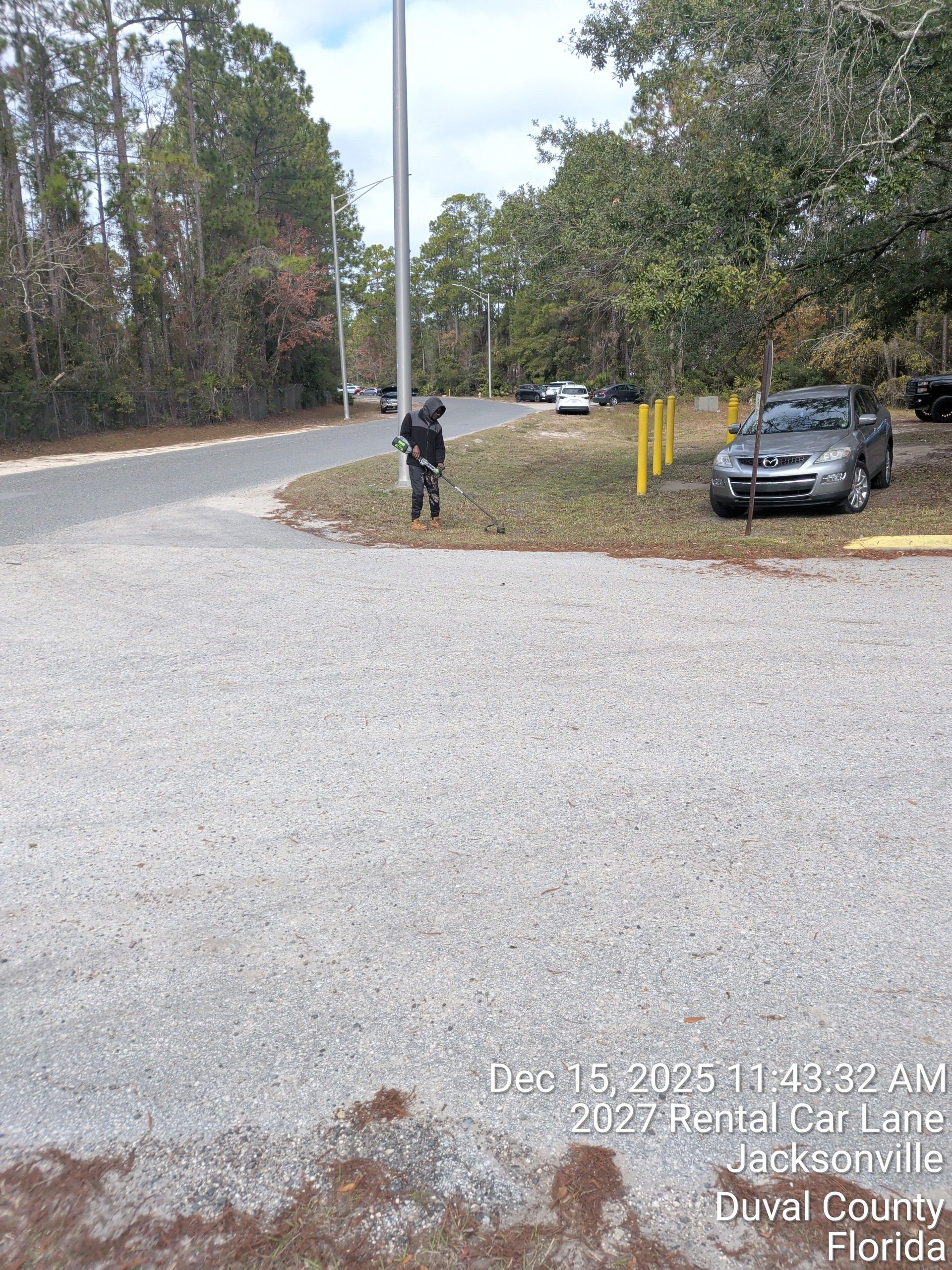 Gravel parking area by a wooded roadside, with a person near a pole and cars parked to the right.