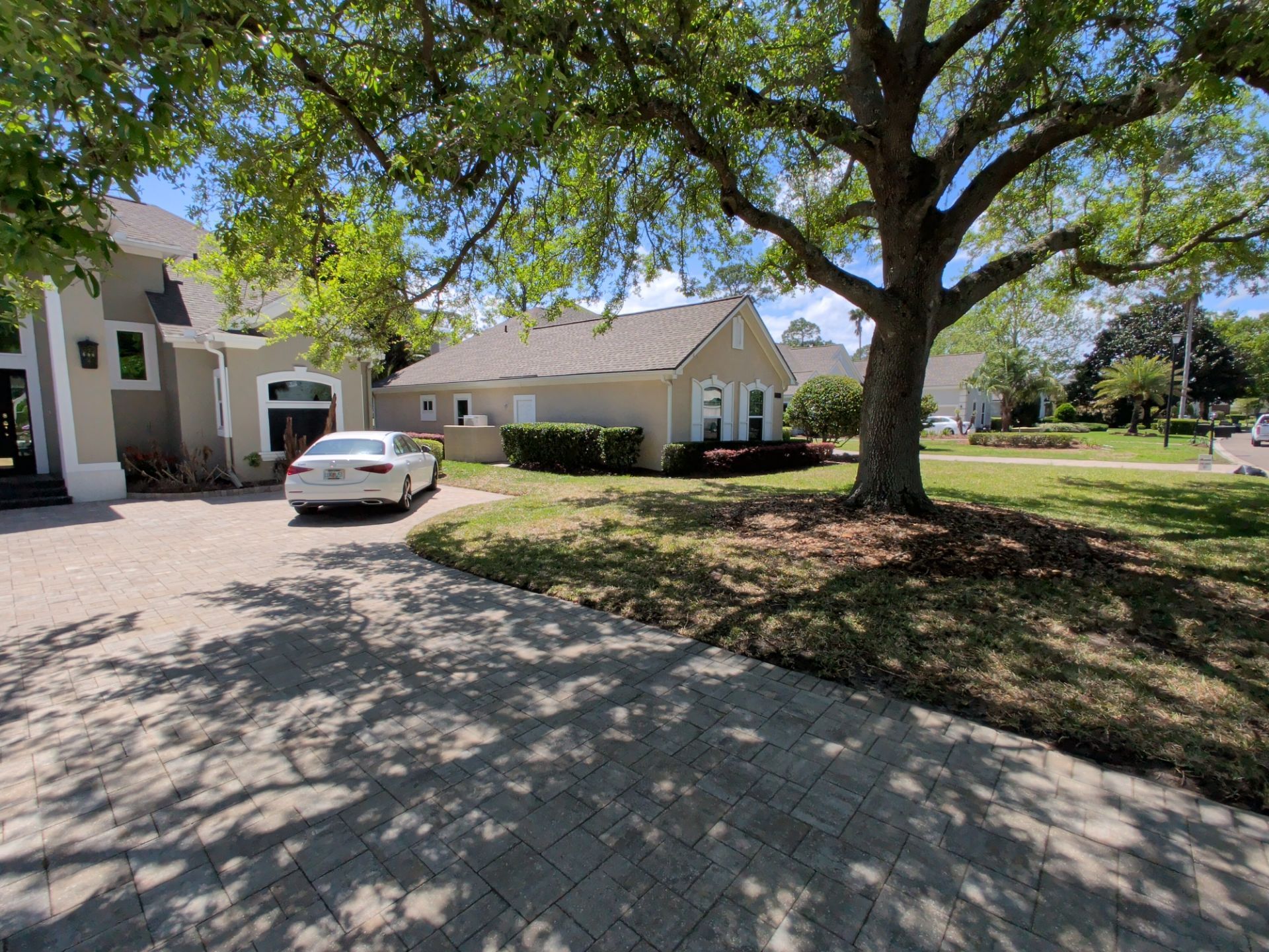Suburban driveway beside a house, with a parked white car and a large tree casting shade.