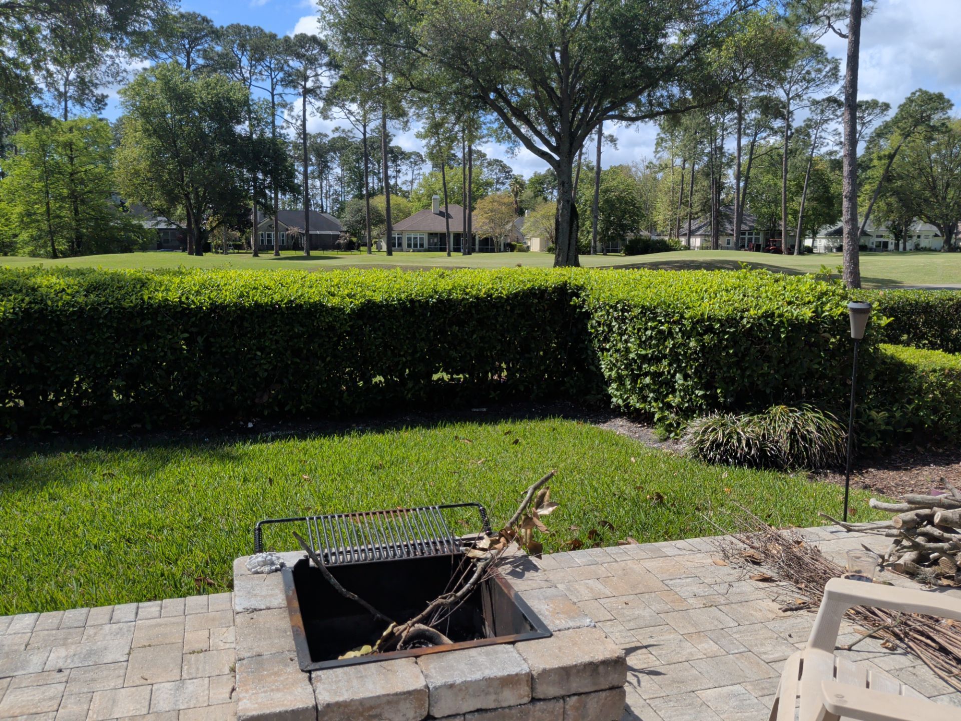 Backyard patio with open fire pit, green lawn, hedge, and trees under a blue sky