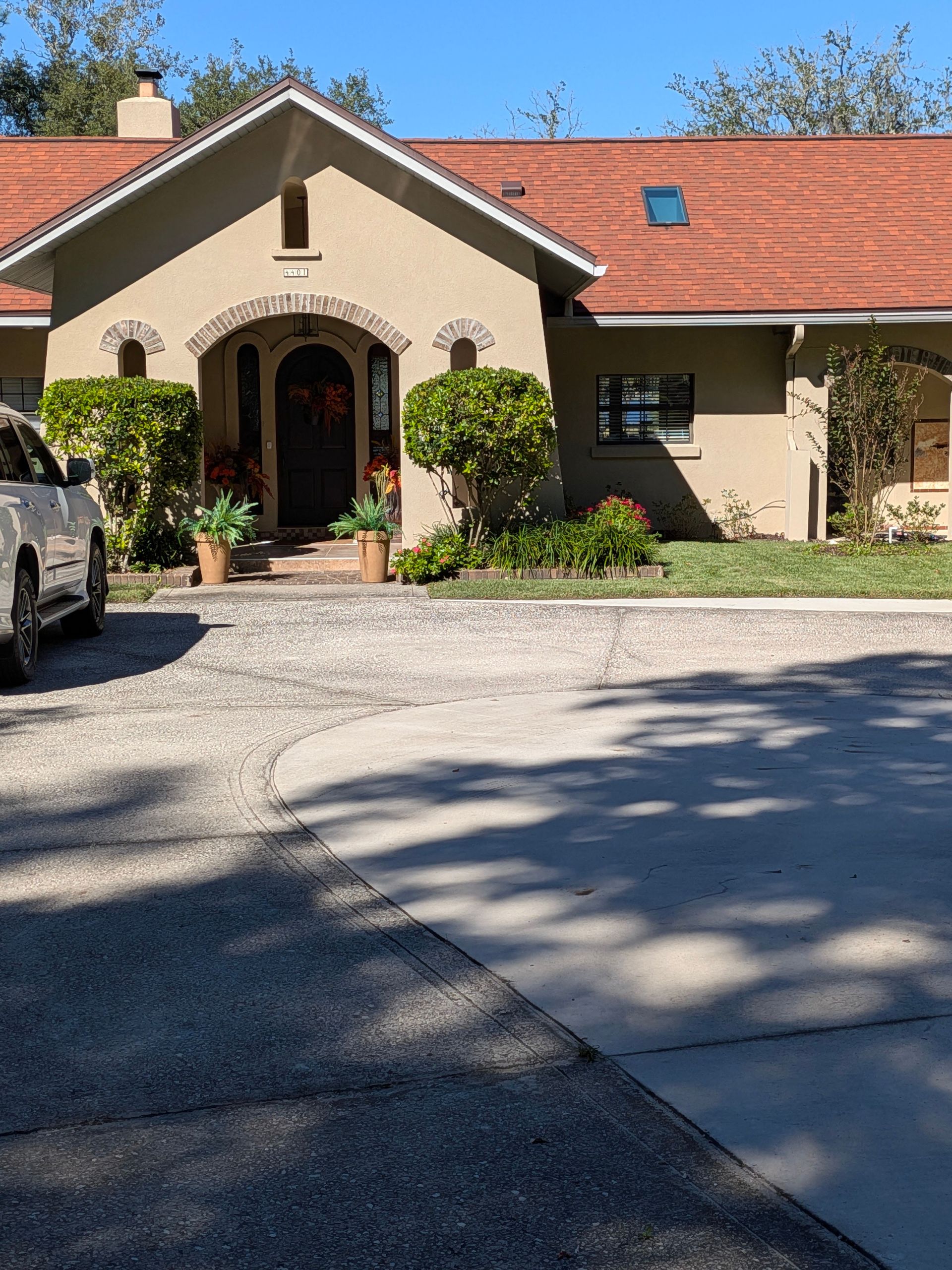 Single-story beige house with red roof, arched front entrance, and gravel driveway in sunlight