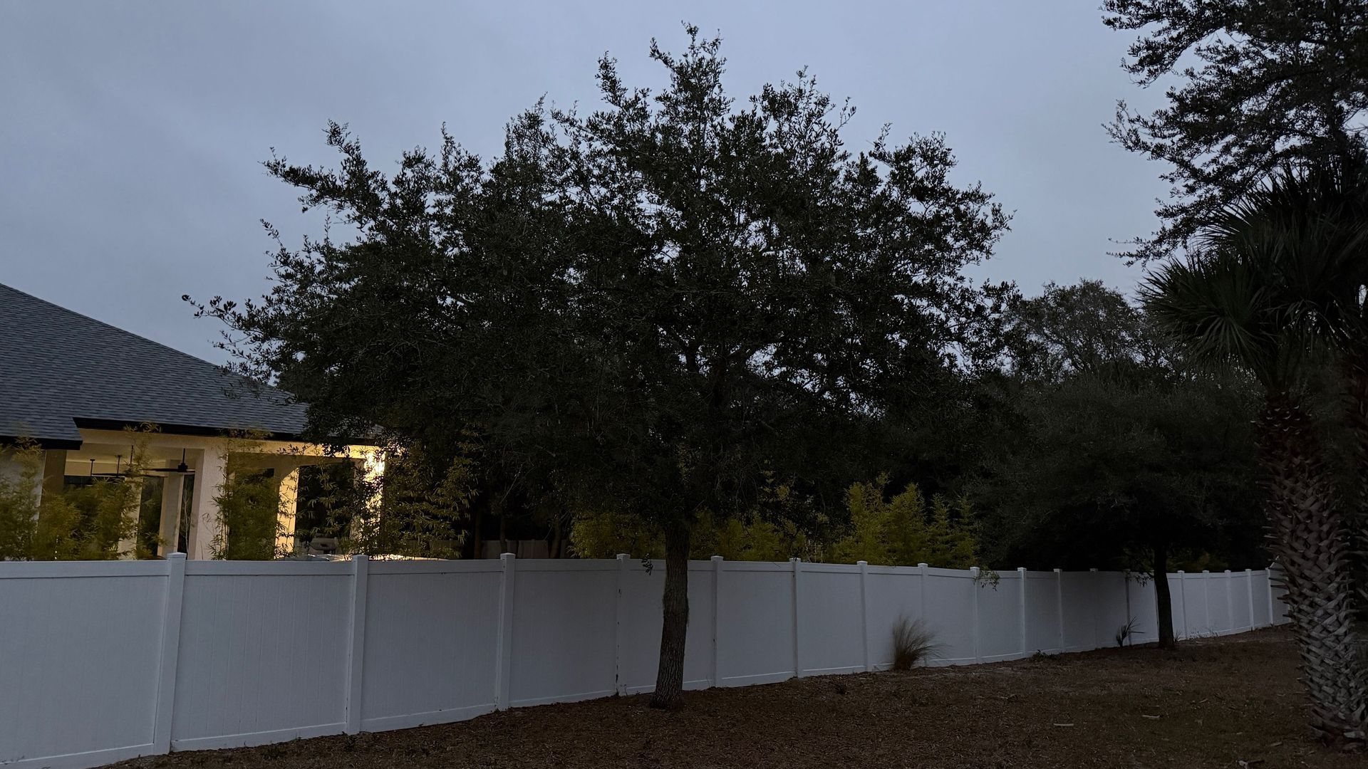 Dusk over a house with lit windows, trees, and a white fence along a driveway.