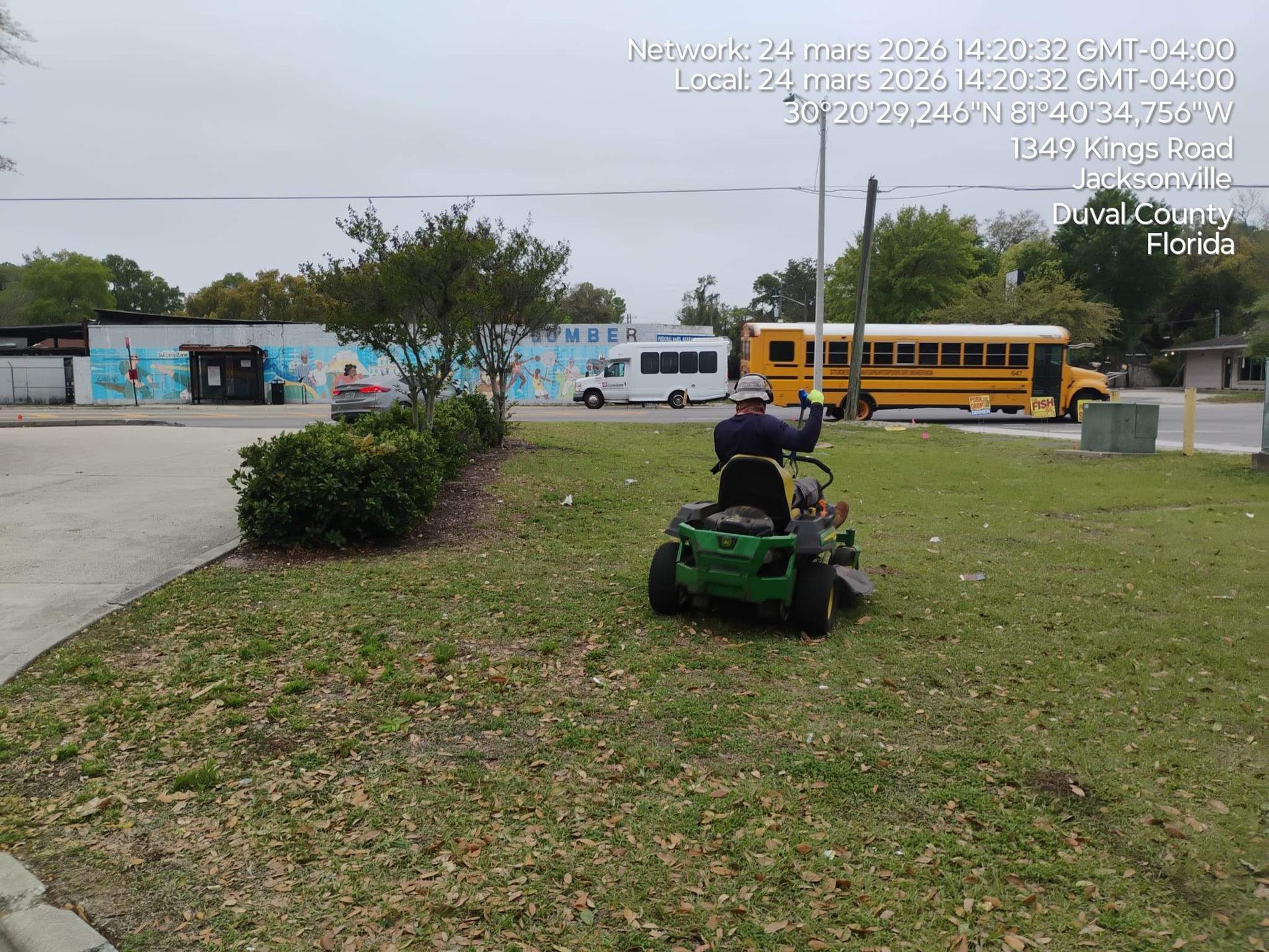 Person on a green lawn mower cutting grass near parked school buses and vans on an overcast day