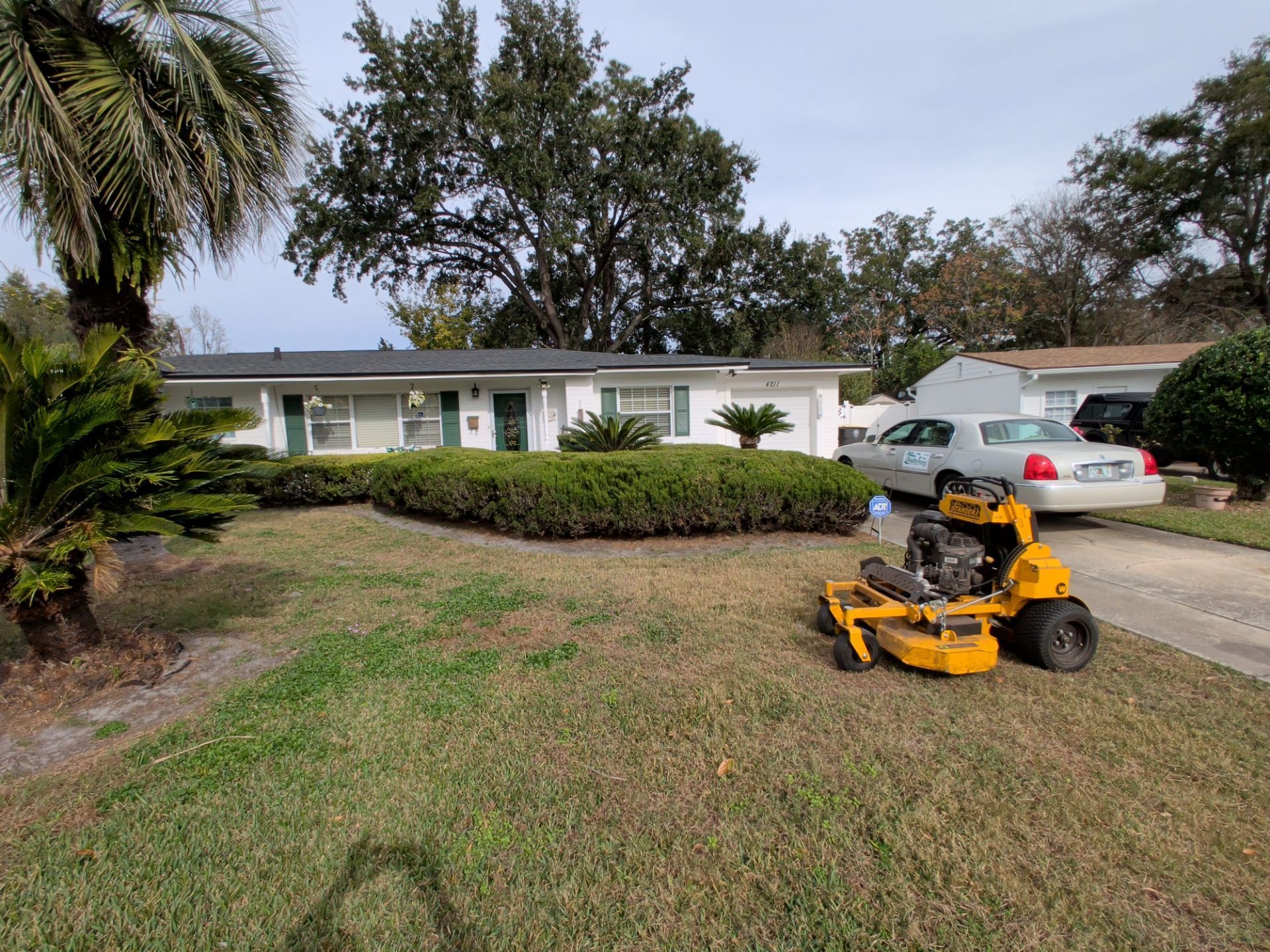 Person mowing a front yard with a yellow riding lawn mower beside a white house and driveway