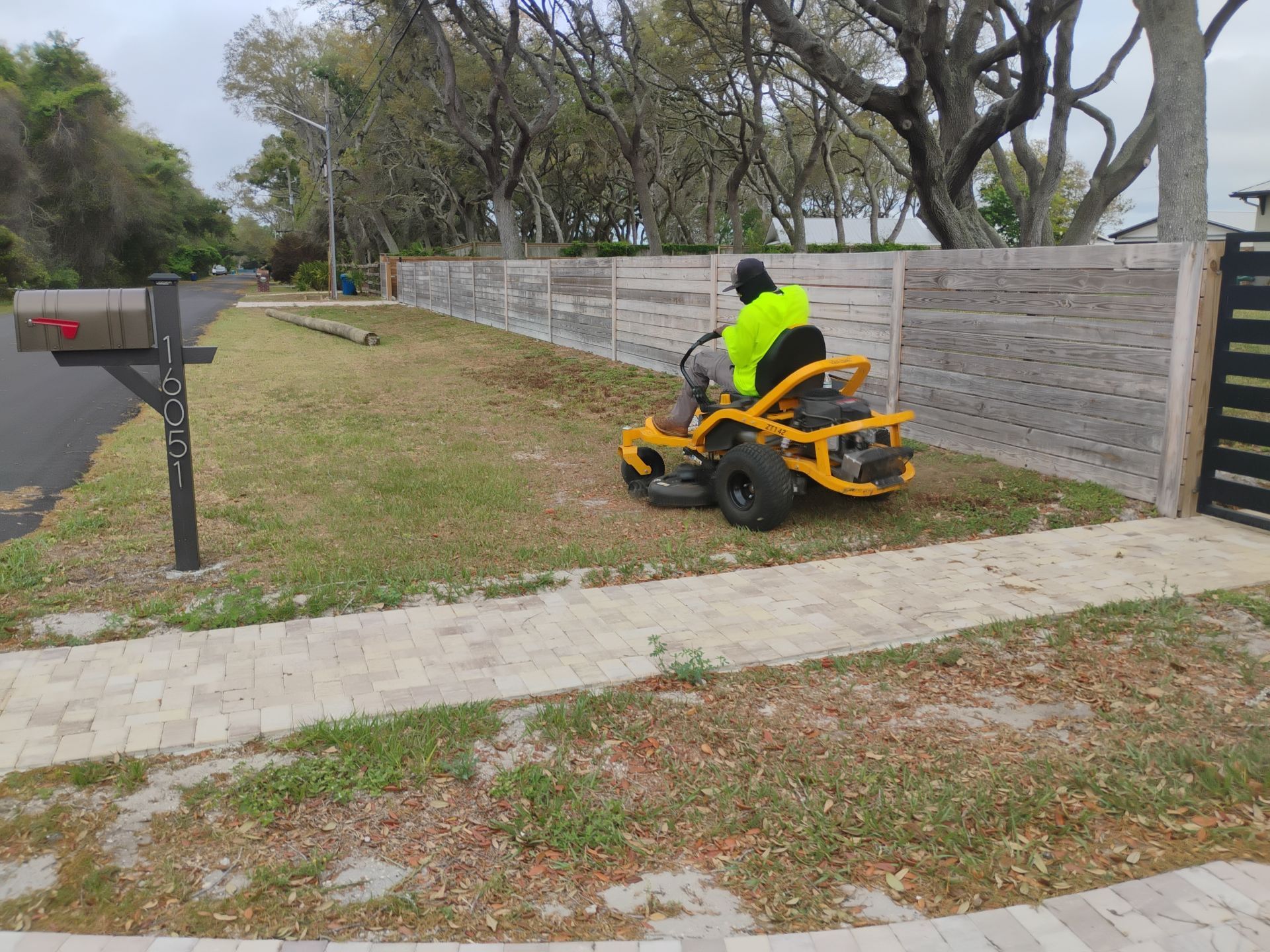 Person in a neon jacket riding a yellow lawn mower along a fenced grassy path near a mailbox.