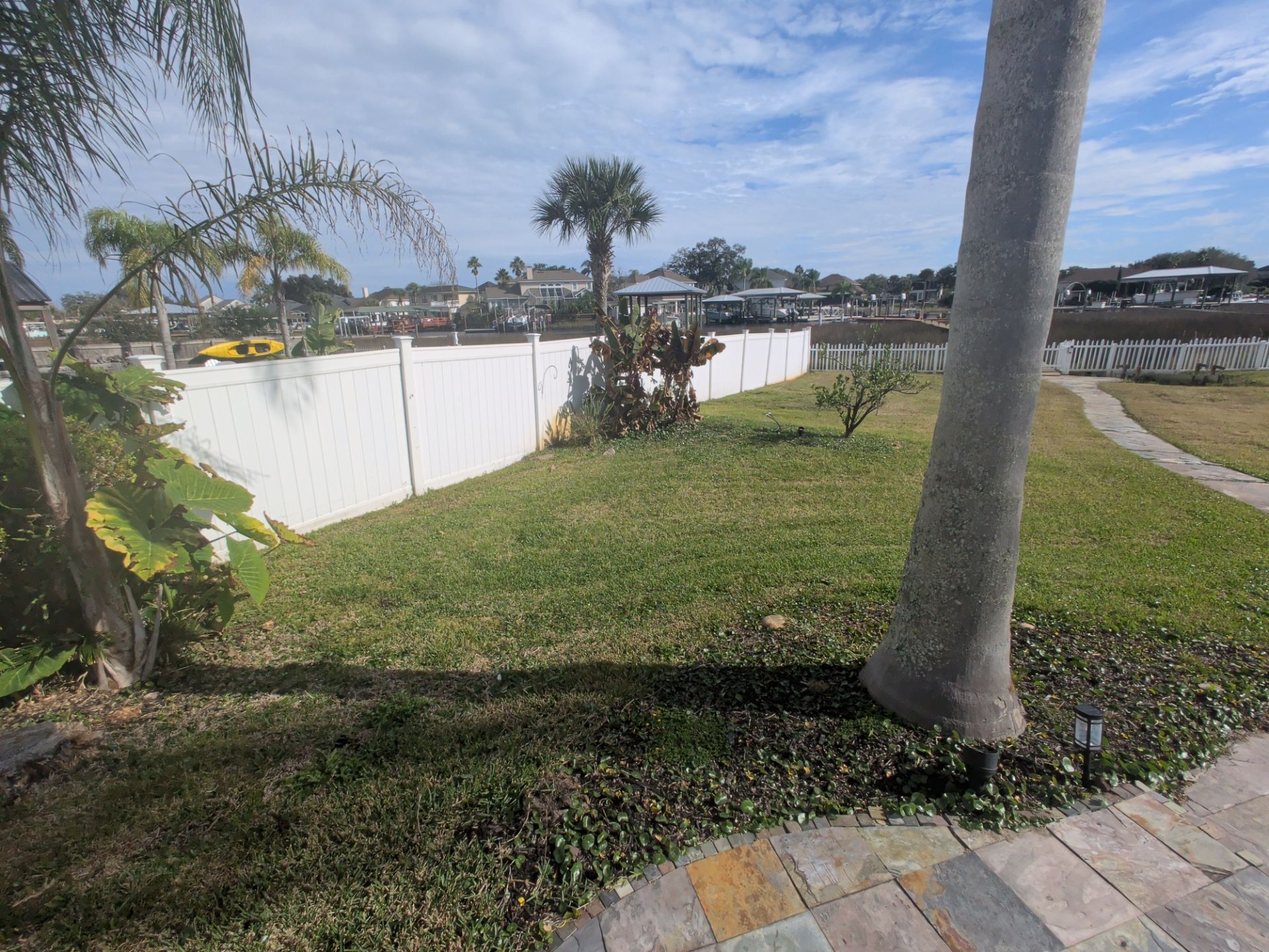 Lawn bordered by white fence, tree in the foreground. Houses and sky visible in the distance.