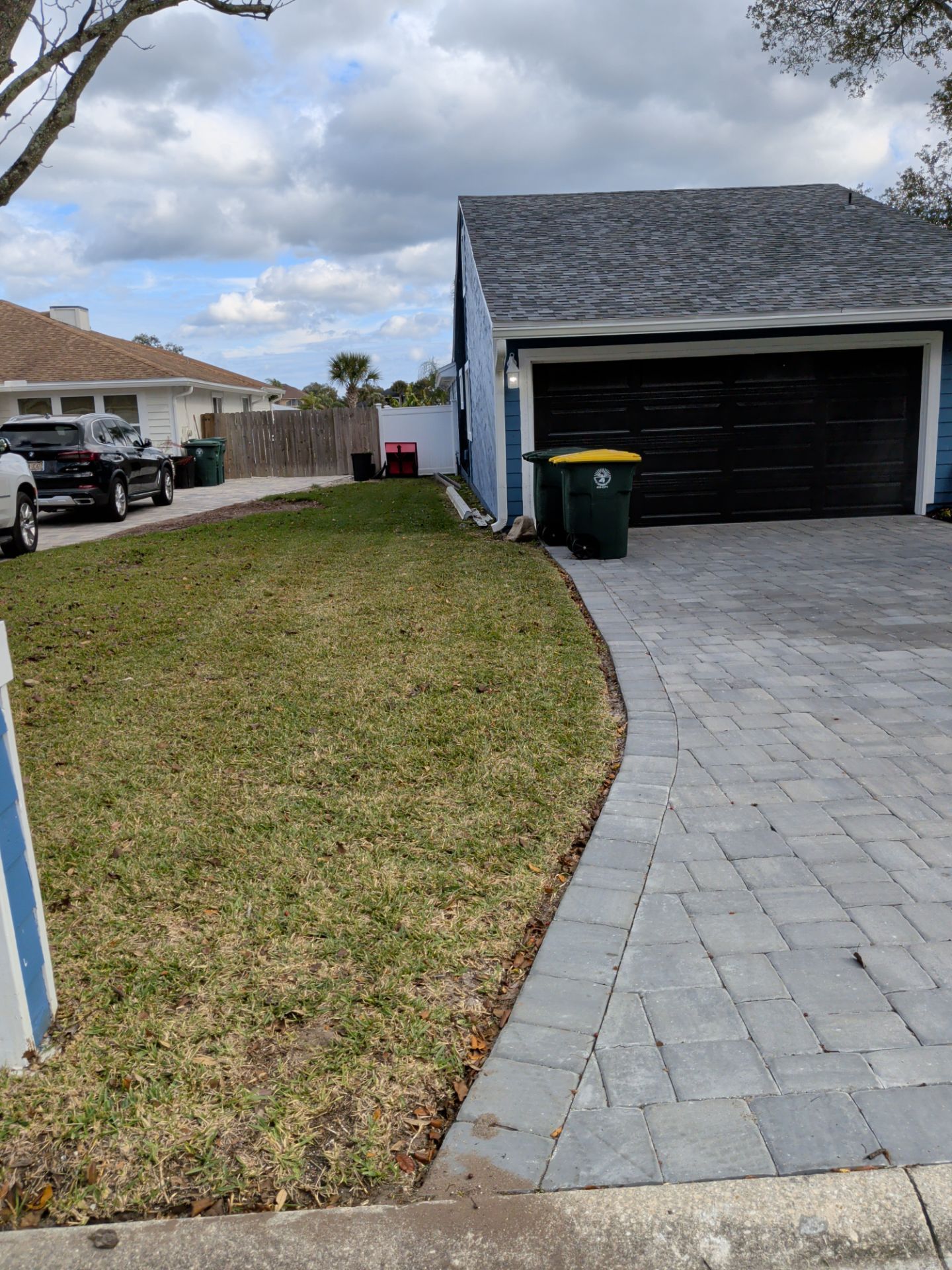 Lawn and paver pathway leading to a blue house with black garage door, green trash bin, and cloudy sky.