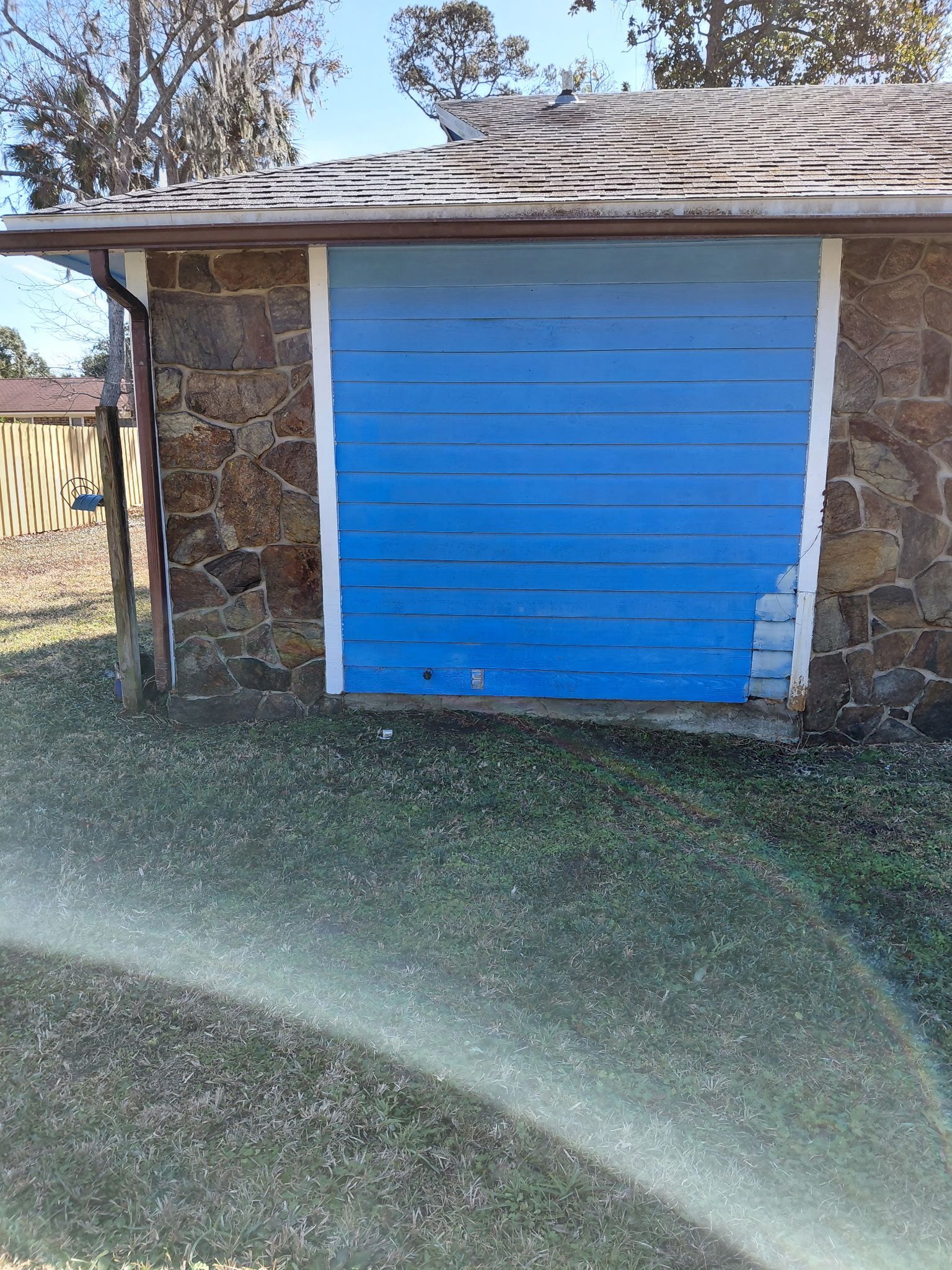 Blue-painted garage door with white trim set into a stone wall, under a roof. Grass and a fence are in the foreground.
