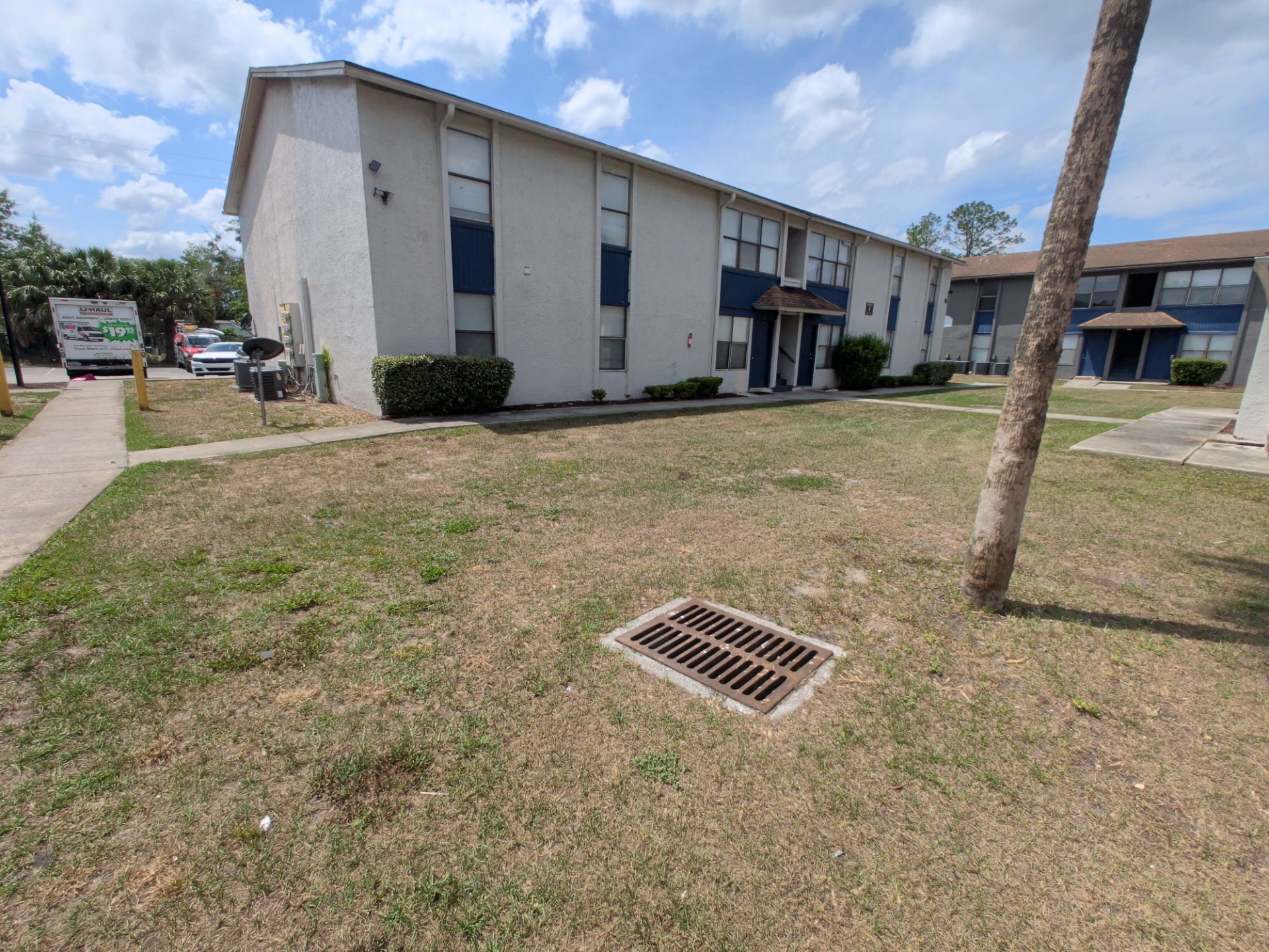 Apartment building with a grassy courtyard, sidewalk, parked cars, and a storm drain under a blue sky