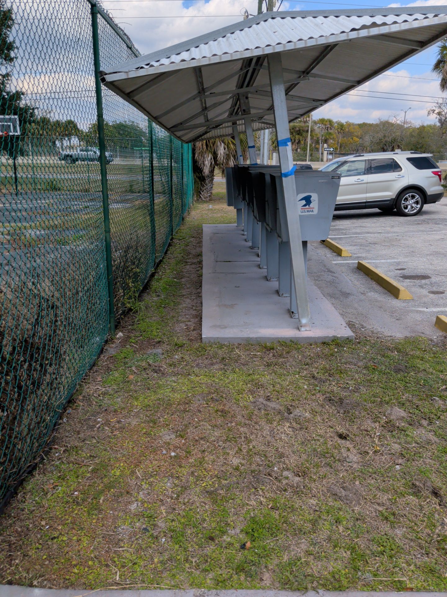 Mailboxes under a covered shelter next to a chain-link fence and parking lot.