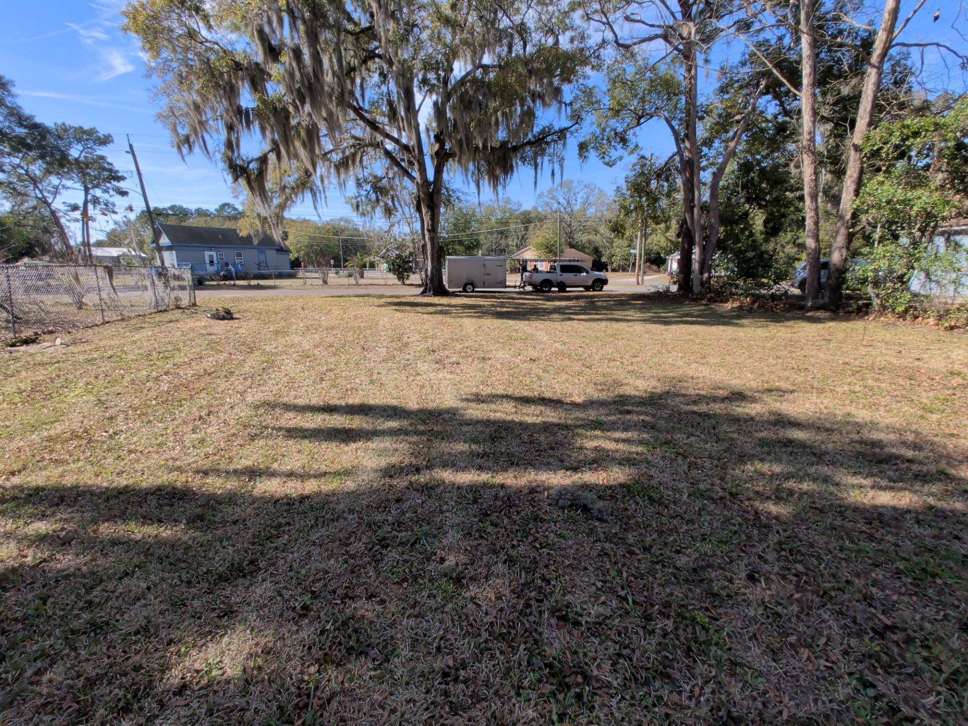 Grassy lot with trees. A vehicle and trailer are parked in the distance; houses are also visible.
