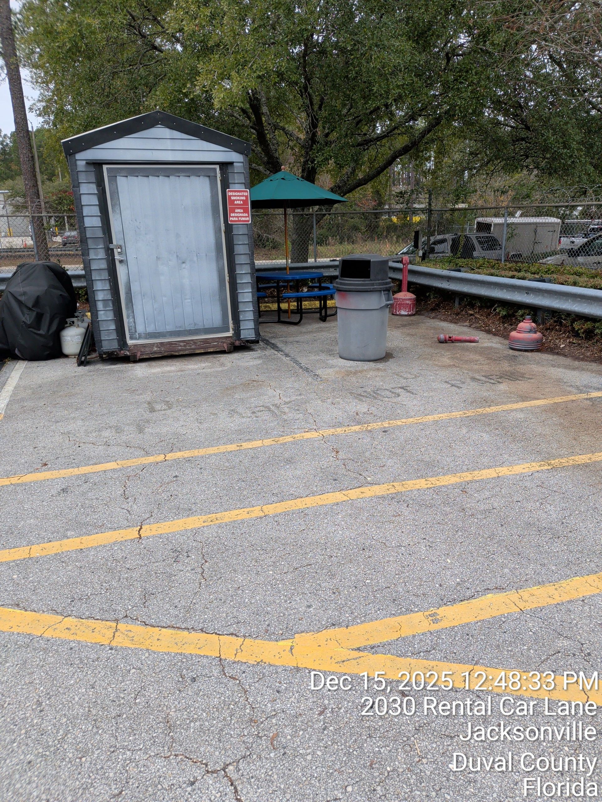 Outdoor parking area with a small shed, trash can, and picnic table under trees.