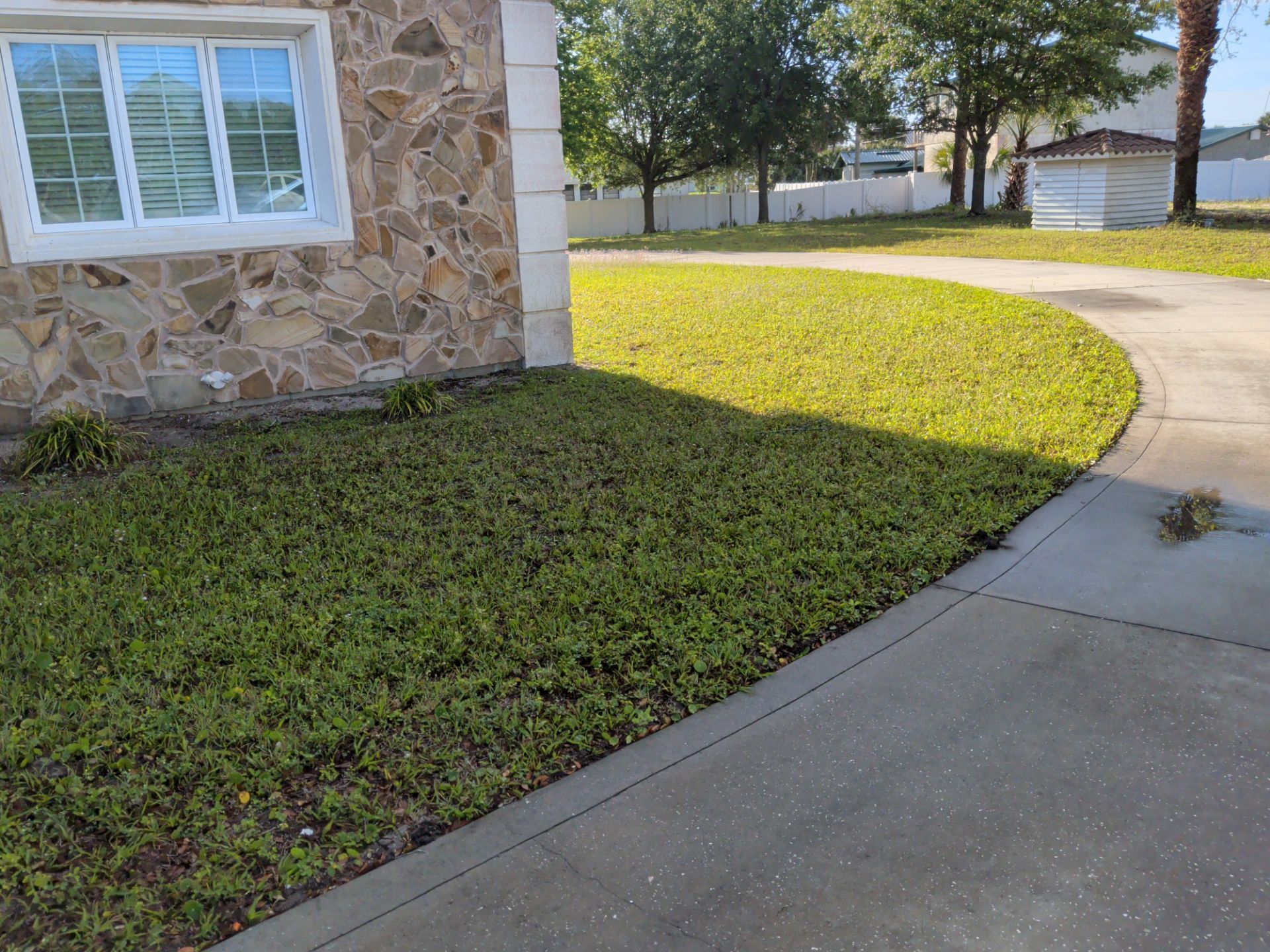 House corner with stone wall, white window, trimmed green lawn, and curved concrete sidewalk in sunlight