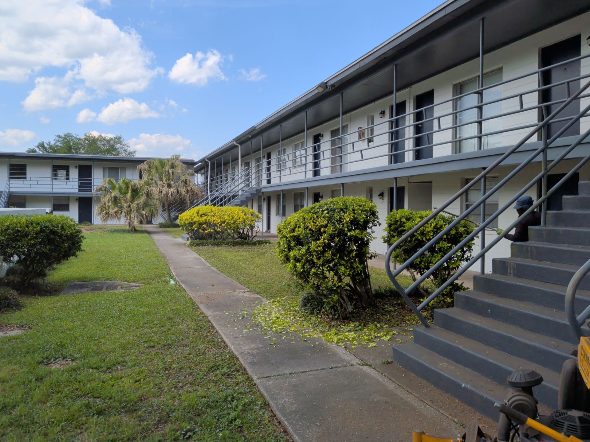 Exterior of a two-story apartment building with stairs, balconies, and a grassy courtyard under a blue sky