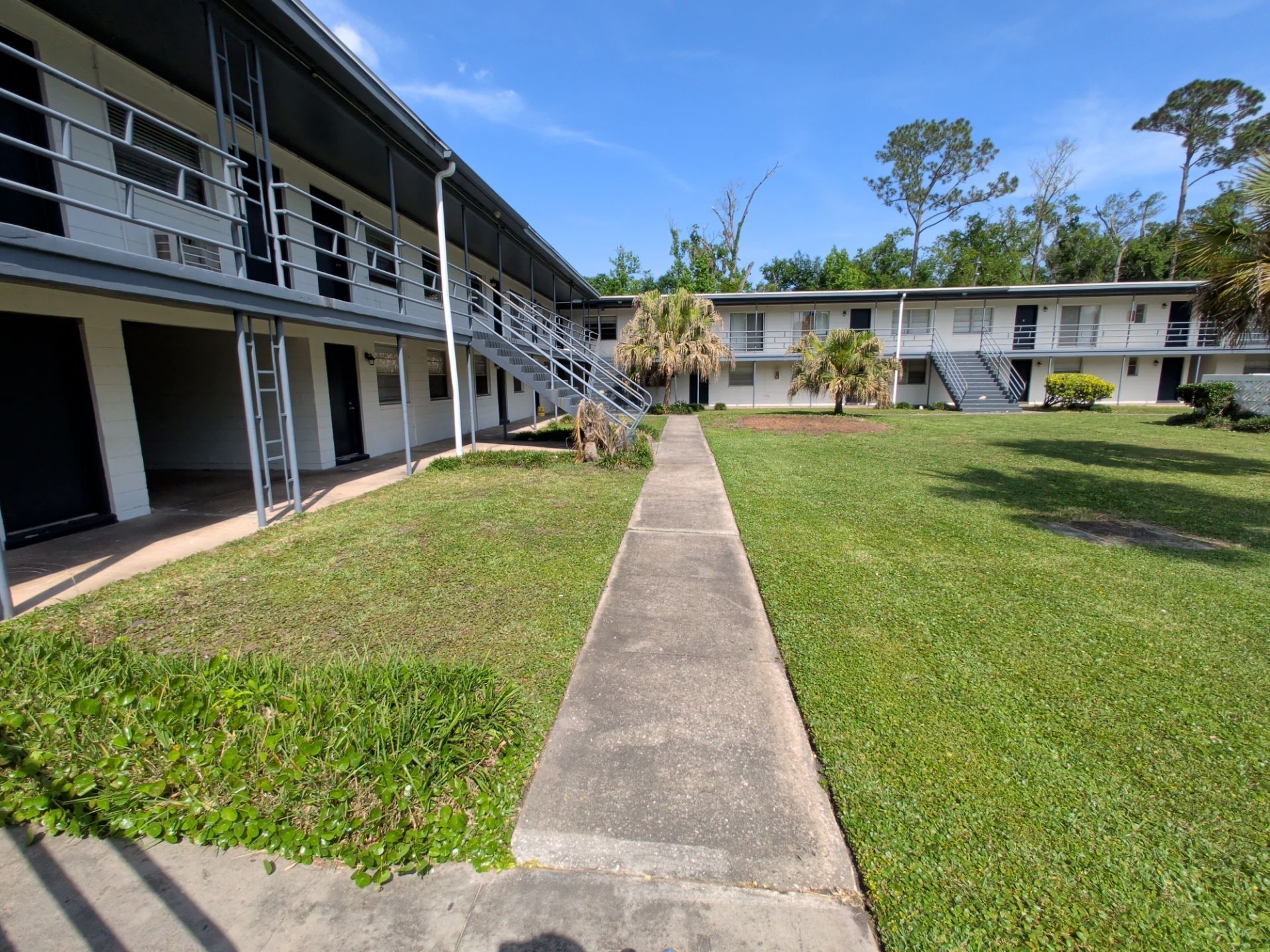 Apartment courtyard with concrete walkway, grassy lawn, and two-story buildings under a sunny sky